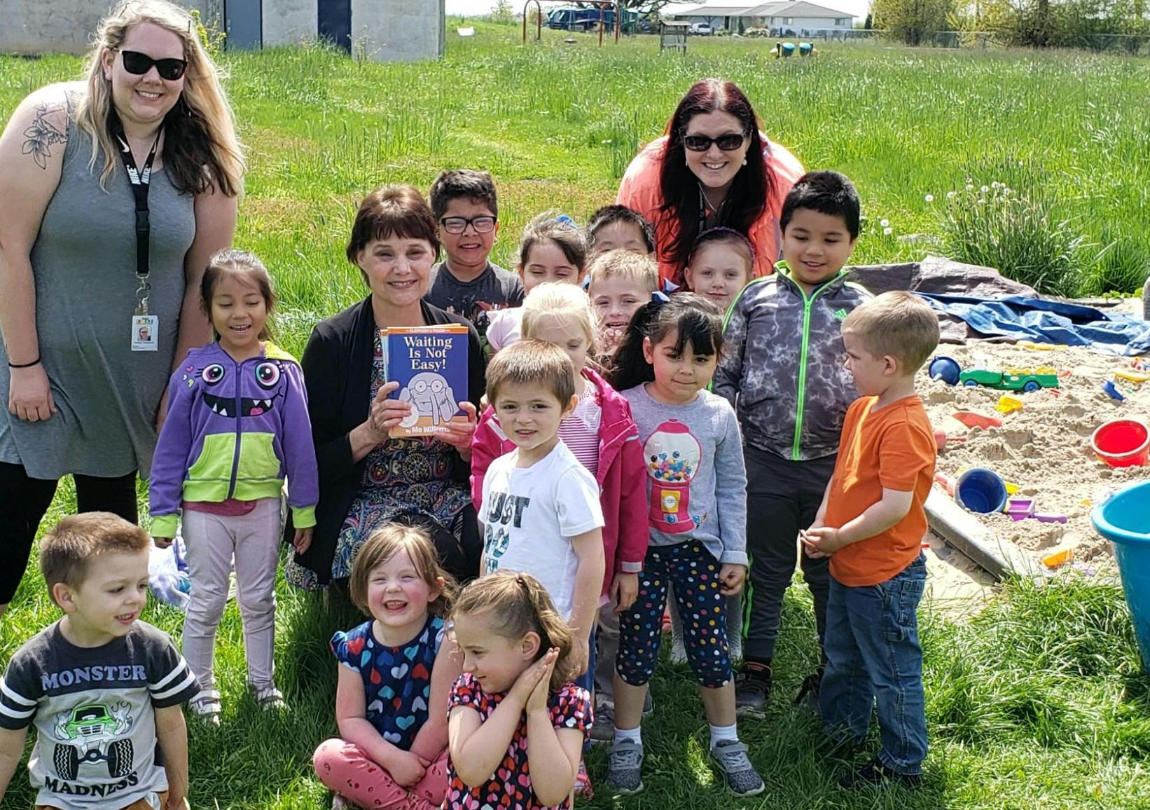Two adults and a group of children outdoors, one adult holding a book