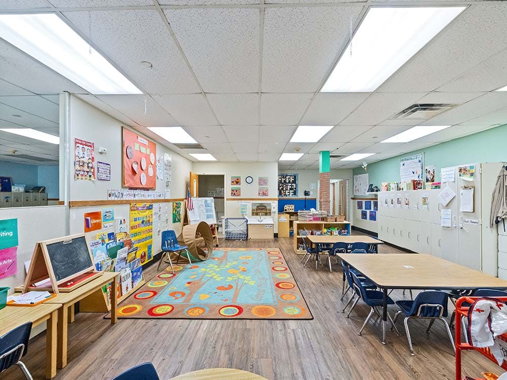 Empty classroom with small tables and chairs, a colorful rug, and cubbies