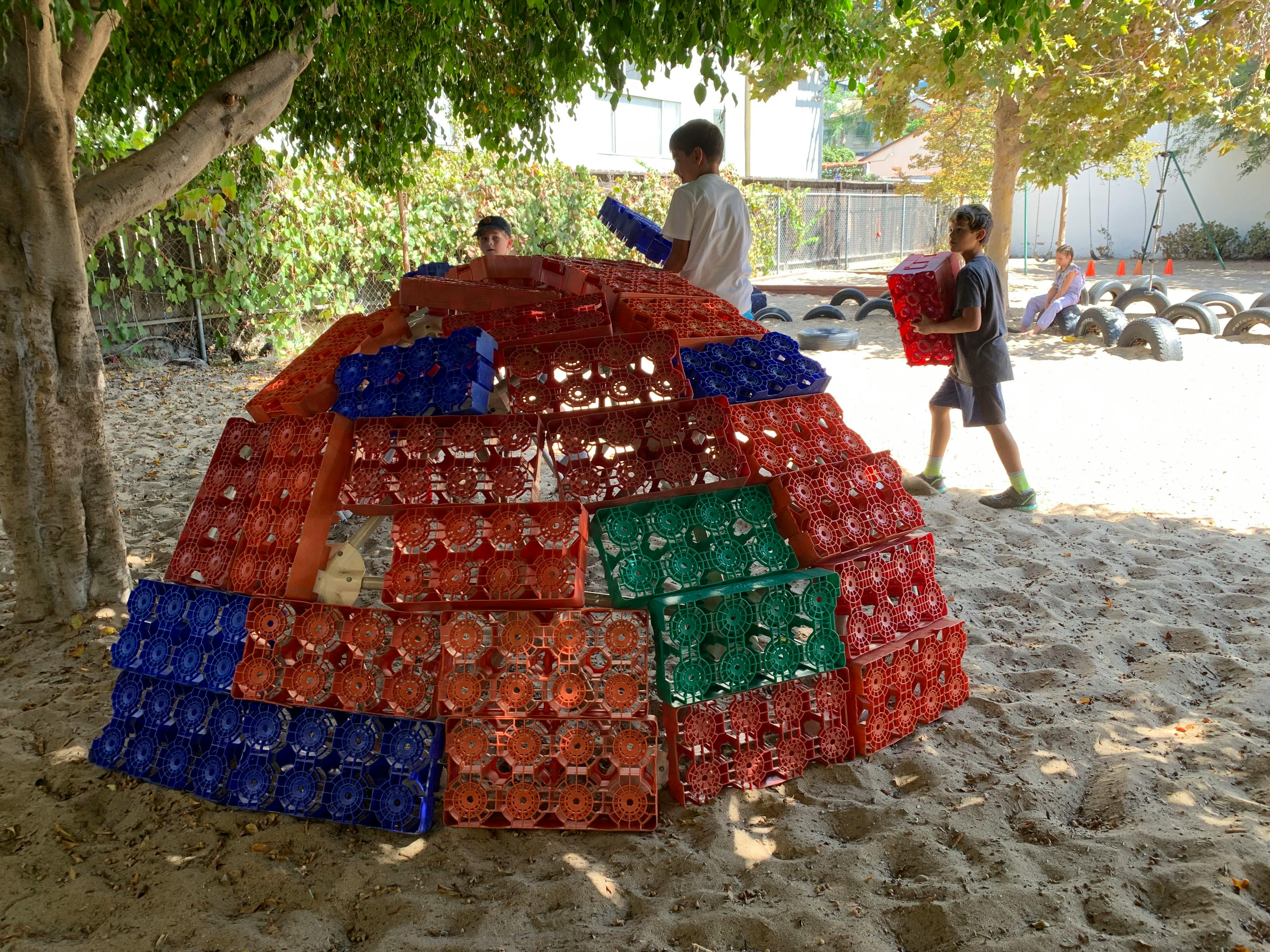 Children building a dome structure from plastic crates in a sandy area