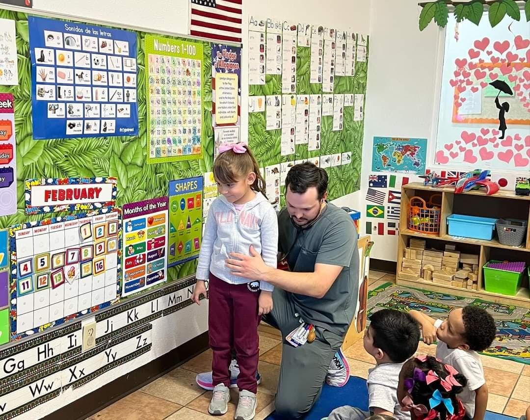 Adult and child standing by a classroom calendar, two children on the floor