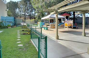 Outdoor playground area with a fence, grass, and covered patio