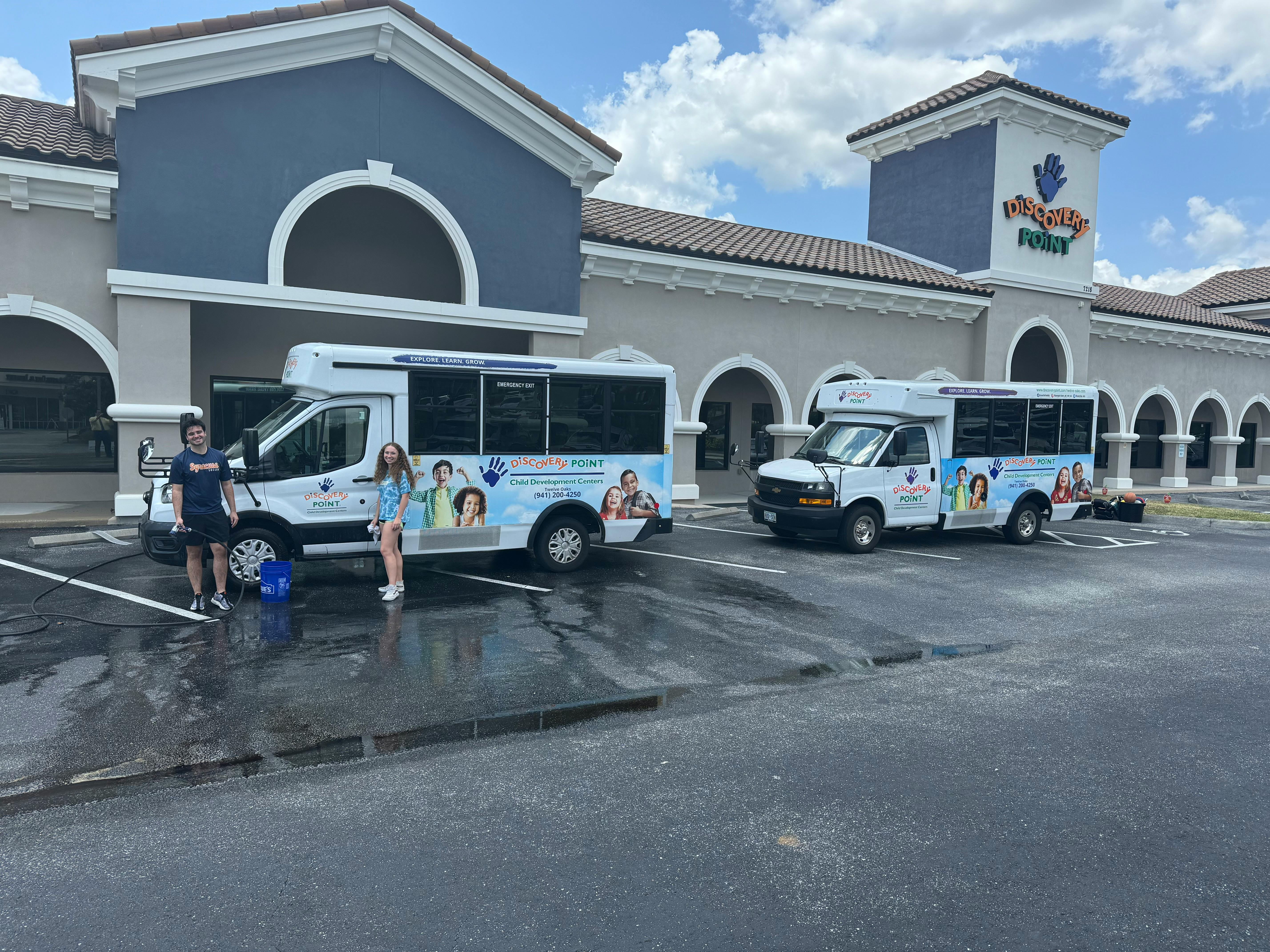 Two people standing next to a white shuttle bus with images of children on its side