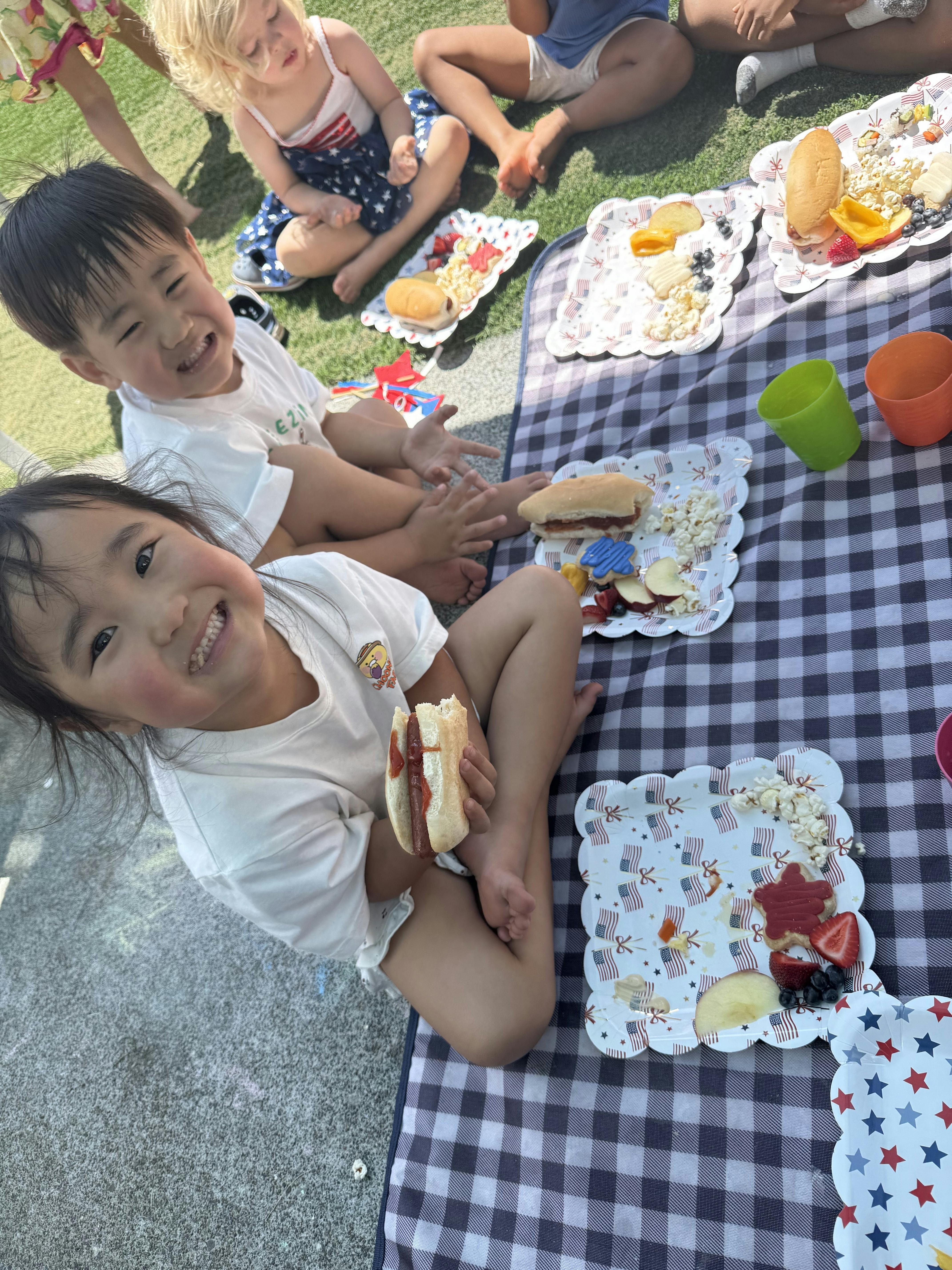 Child holding a hot dog, with other children and picnic food on a blanket
