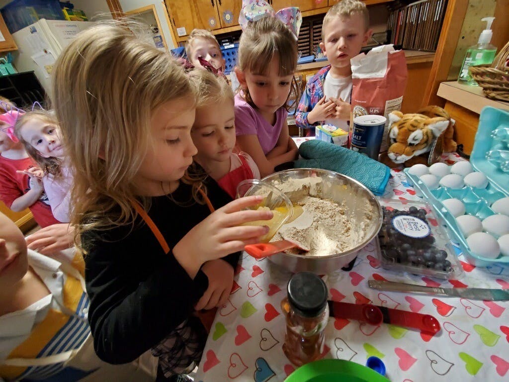 Children gathered around a table, one child pouring liquid into a bowl of batter