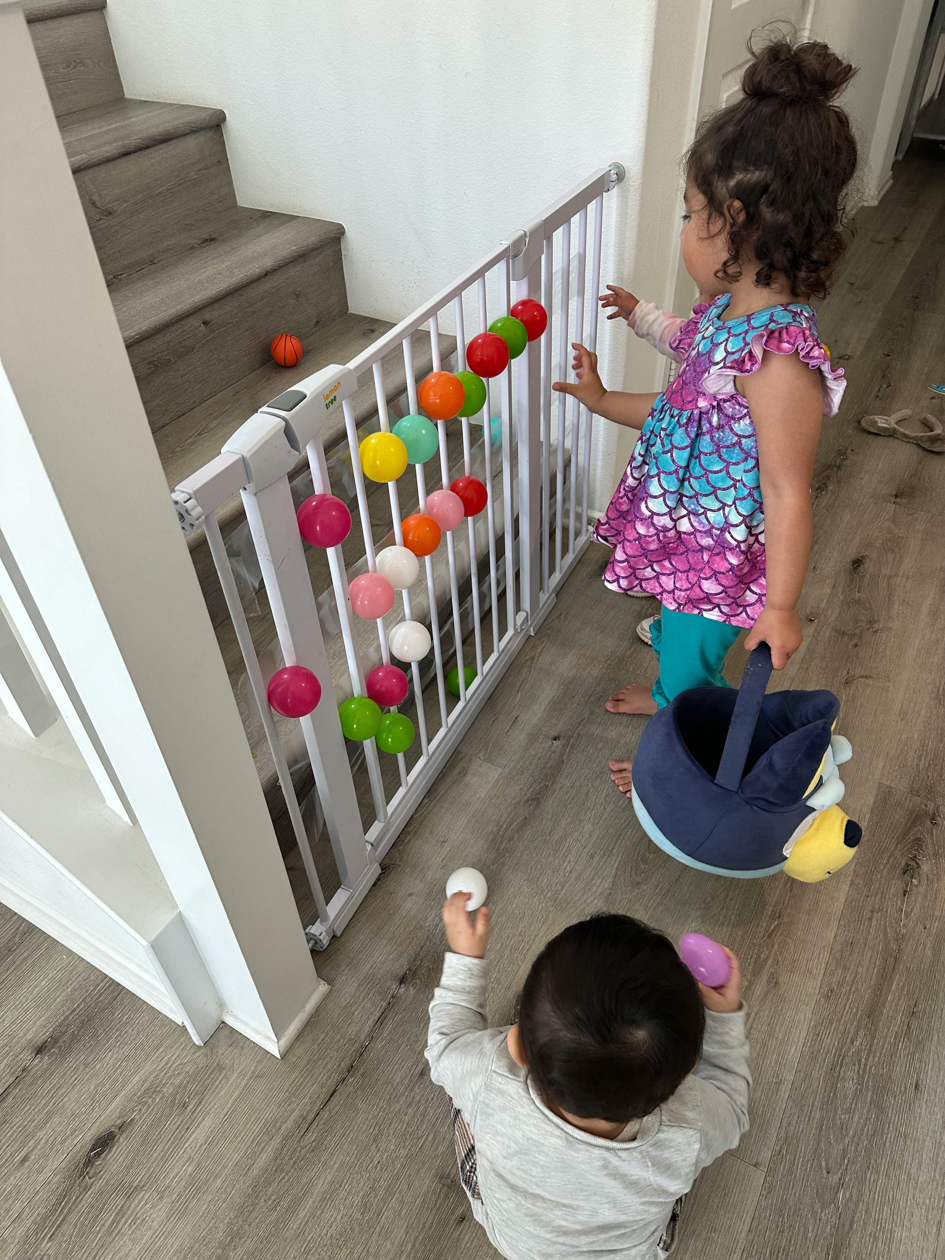 Child standing by a baby gate with colorful balls, another child on the floor