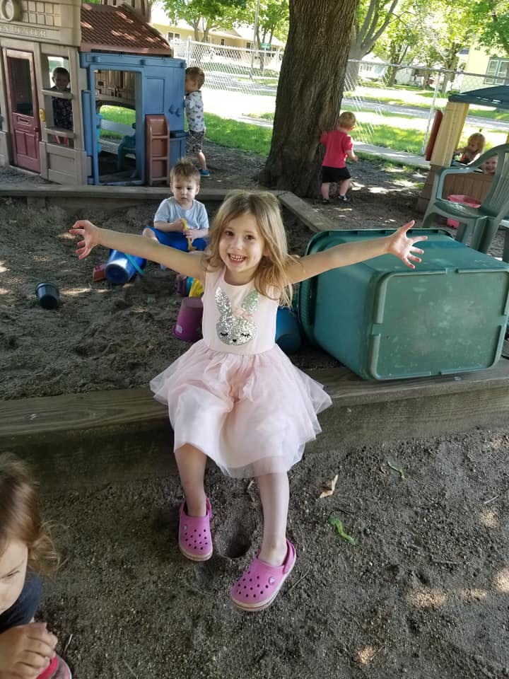 Child in pink dress with arms outstretched, sitting in a sandbox