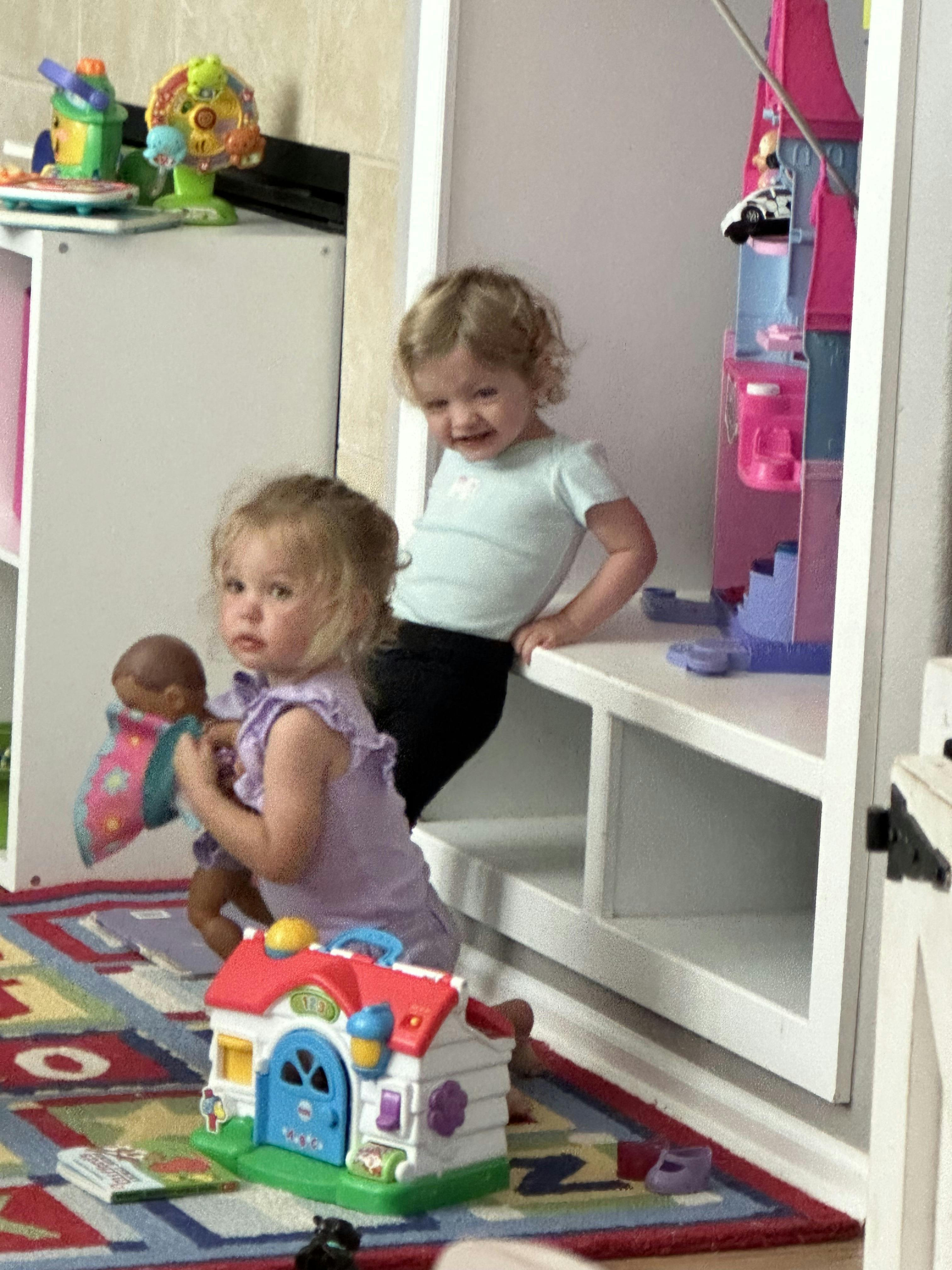 Two children playing with toys, one holding a doll and the other standing by a shelf