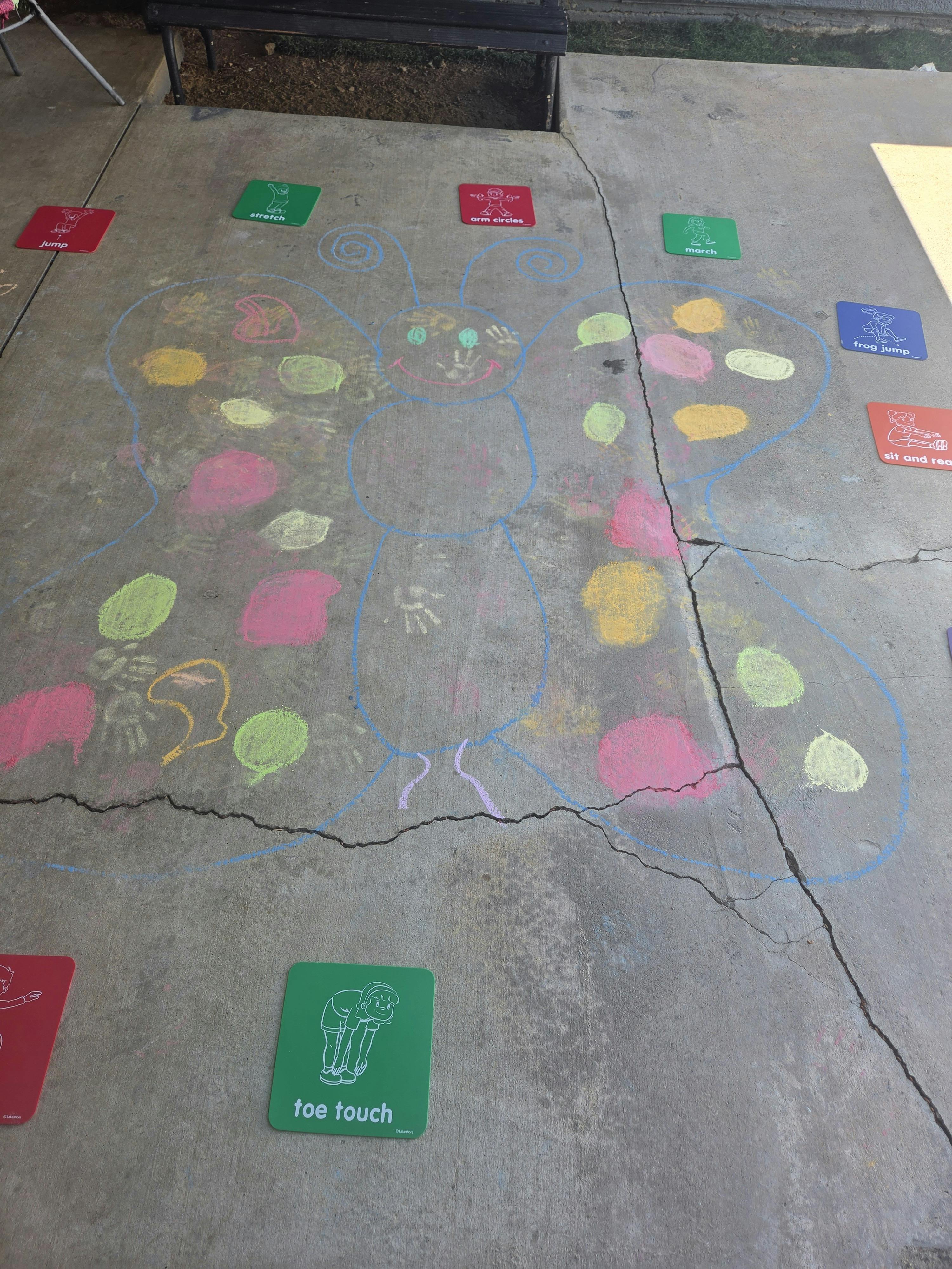 Chalk drawing of a butterfly on cracked concrete, with activity cards for 'jump', 'stretch', 'arm circles', 'march', 'frog jump', 'sit and read', and 'toe touch' laid around it