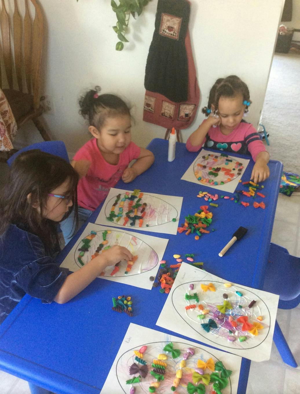 Three children sitting at a blue table making pasta art