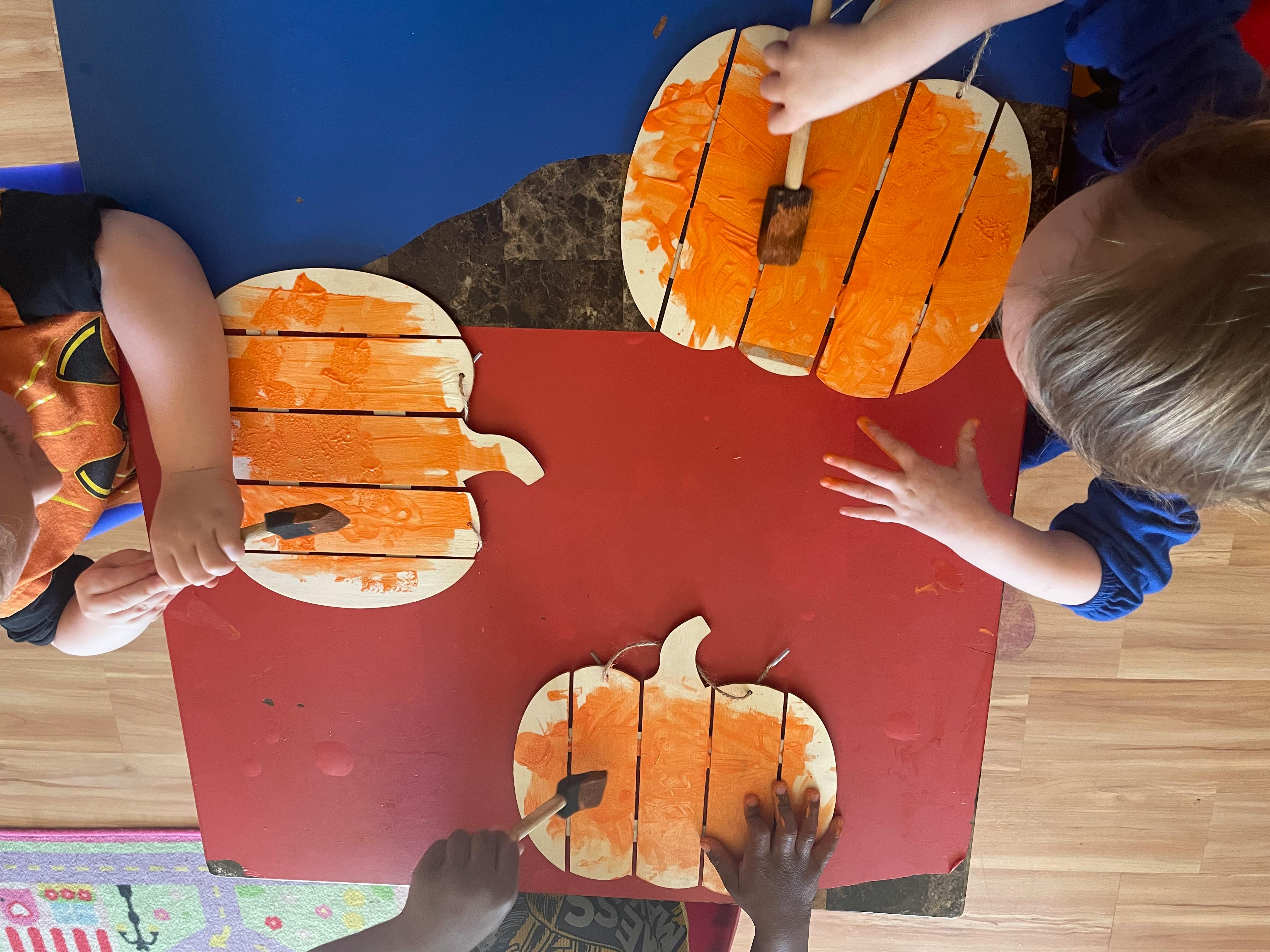 Children painting wooden pumpkin cutouts orange at a table