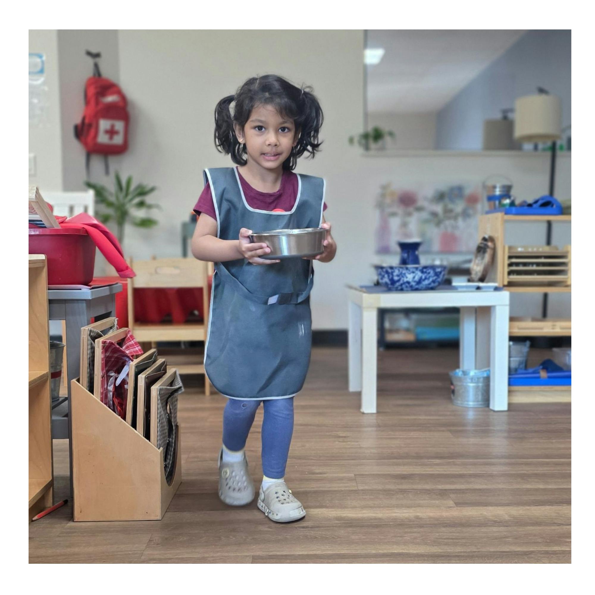 Child wearing an apron holding a small metal bowl in a classroom