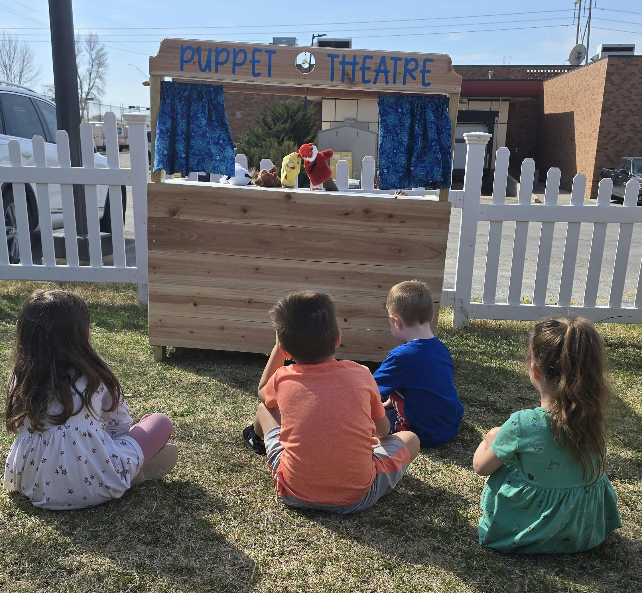 Children sitting on grass watching a puppet show at a puppet theatre