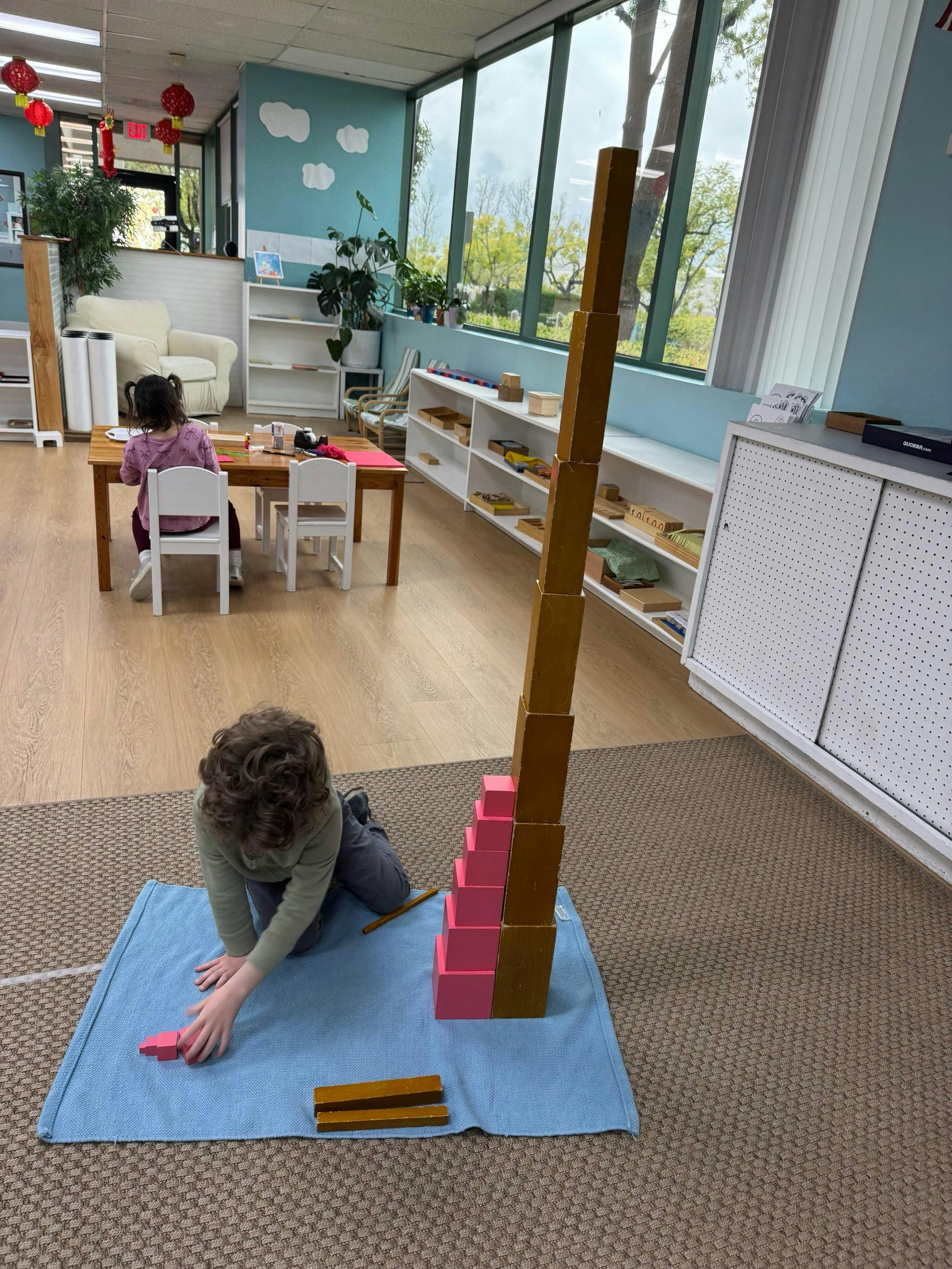 Child building with blocks on the floor, another child sitting at a table