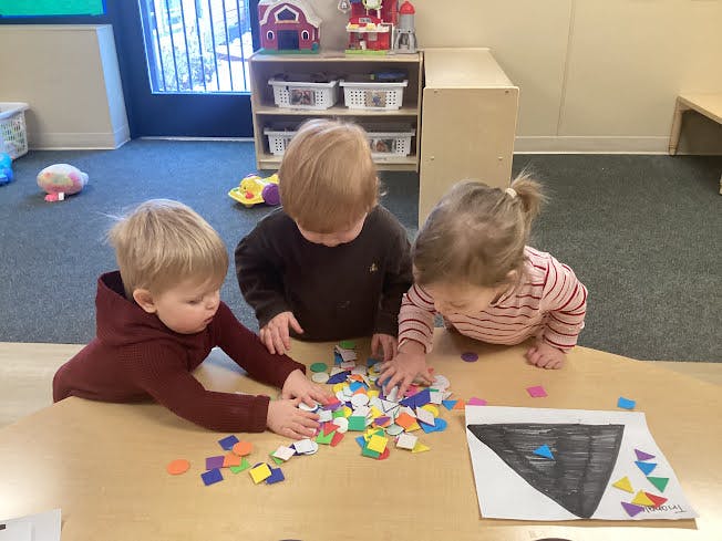 Three children playing with colorful foam shapes on a table in a classroom