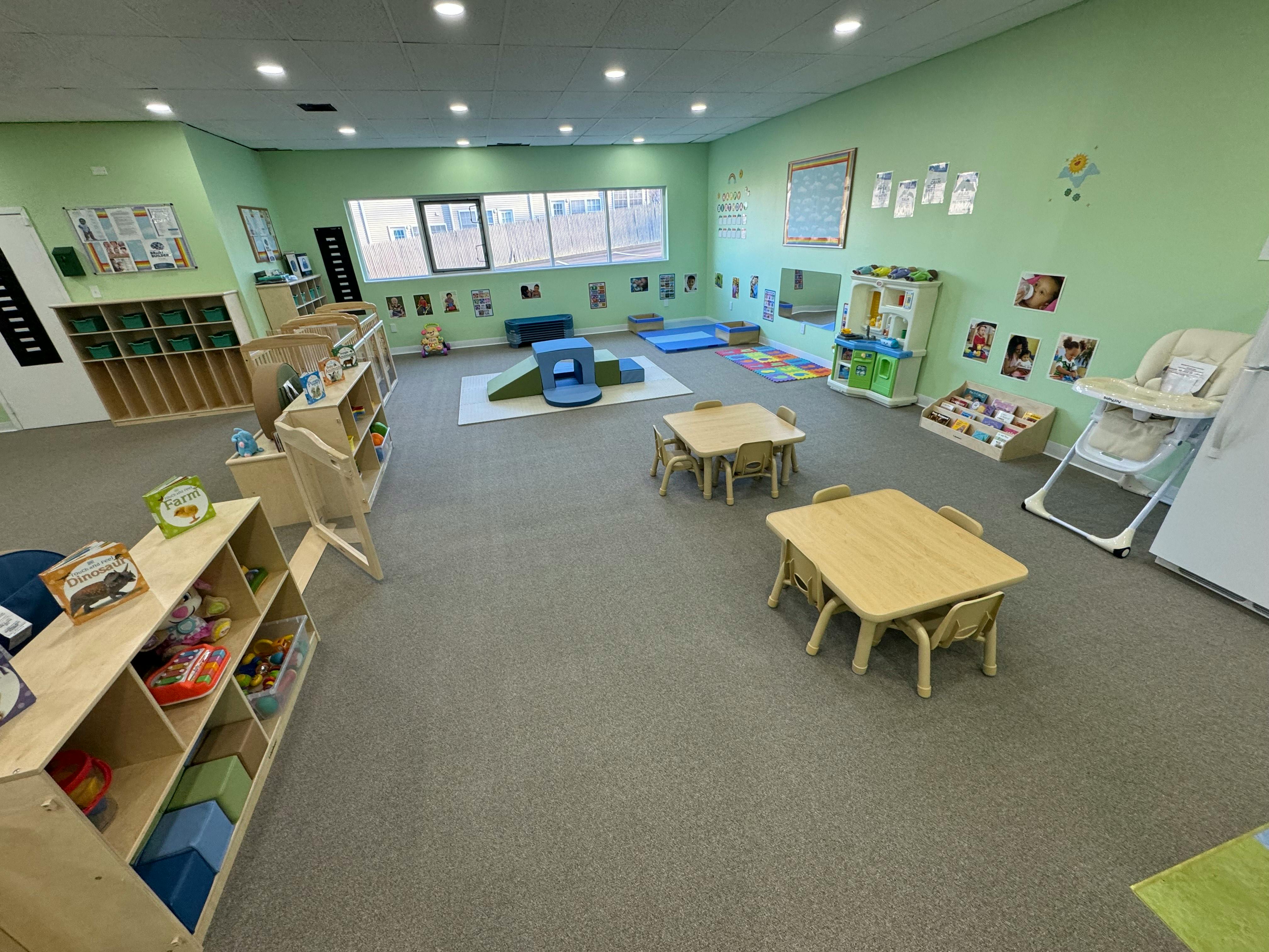 Childcare room with small tables, play kitchen, soft play area, and cribs