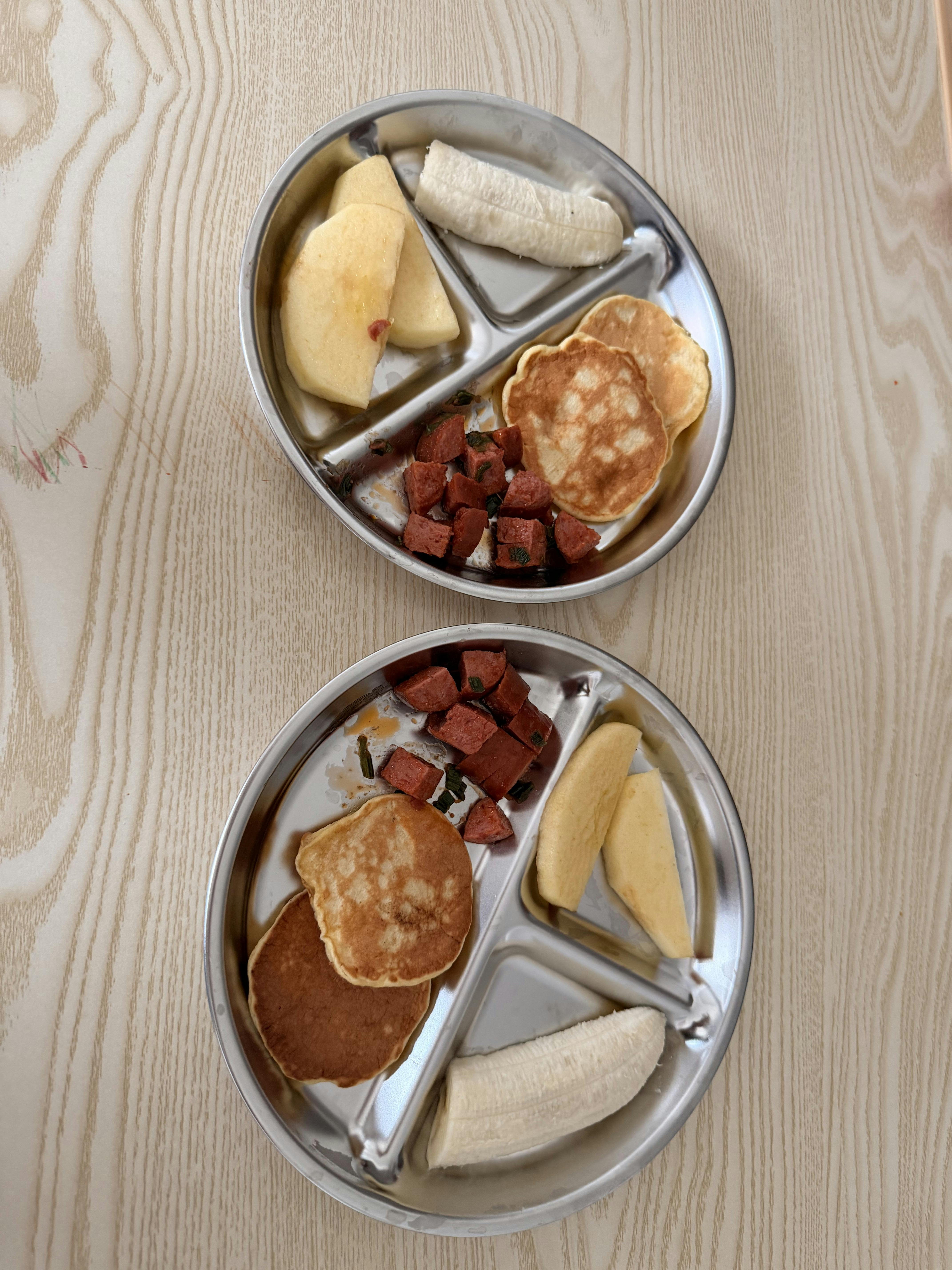 Two divided stainless steel plates with pancakes sliced sausage and fruit