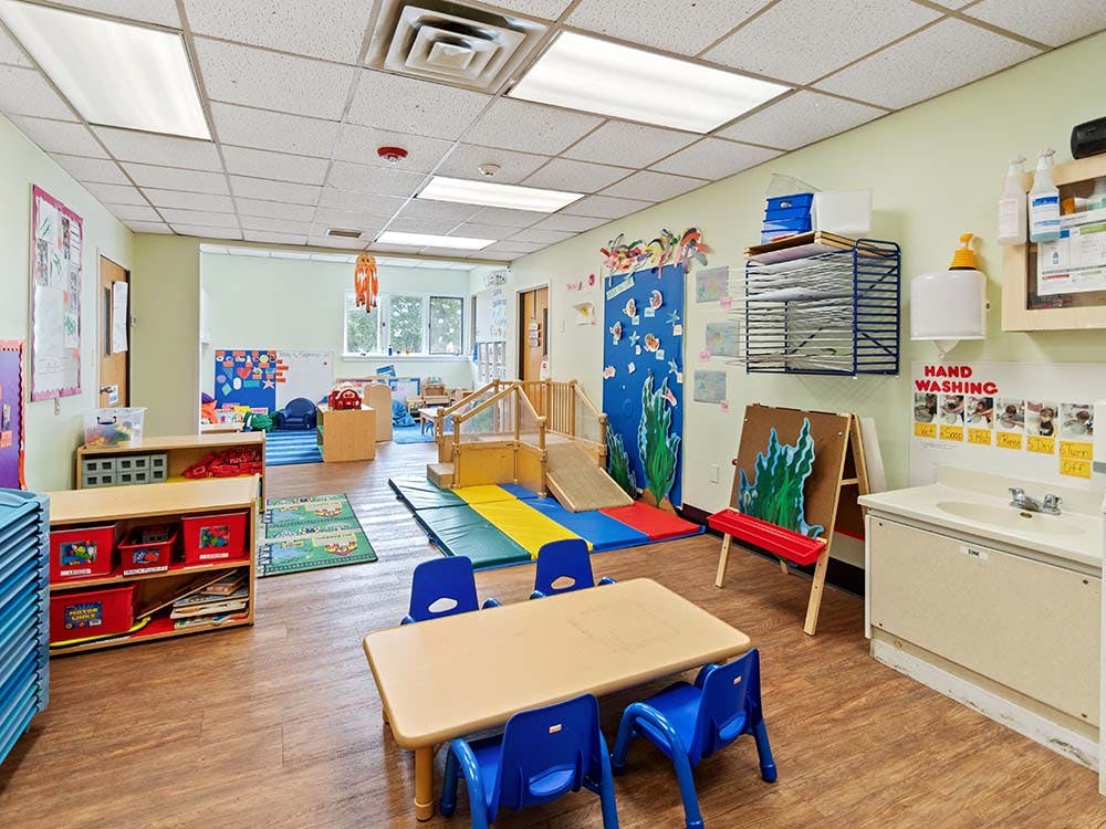 An empty classroom with small tables and chairs, play mats, and a handwashing station