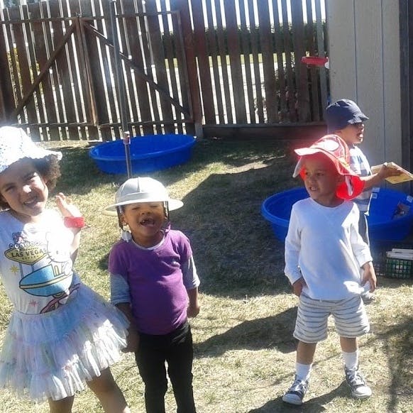 Three children playing outdoors near a wooden fence and blue tubs