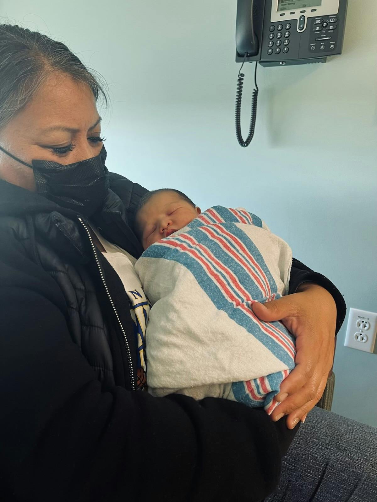 Person wearing a face mask holding a newborn child wrapped in a striped blanket