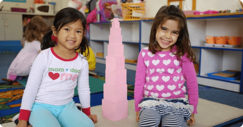 Two children sitting next to a pink conical tower in a classroom
