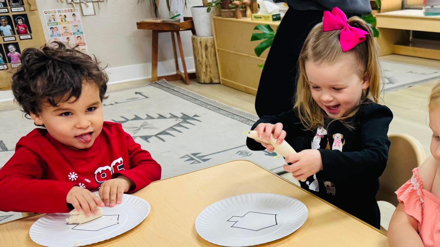 Two children at a table, one making a silly face, the other holding dough