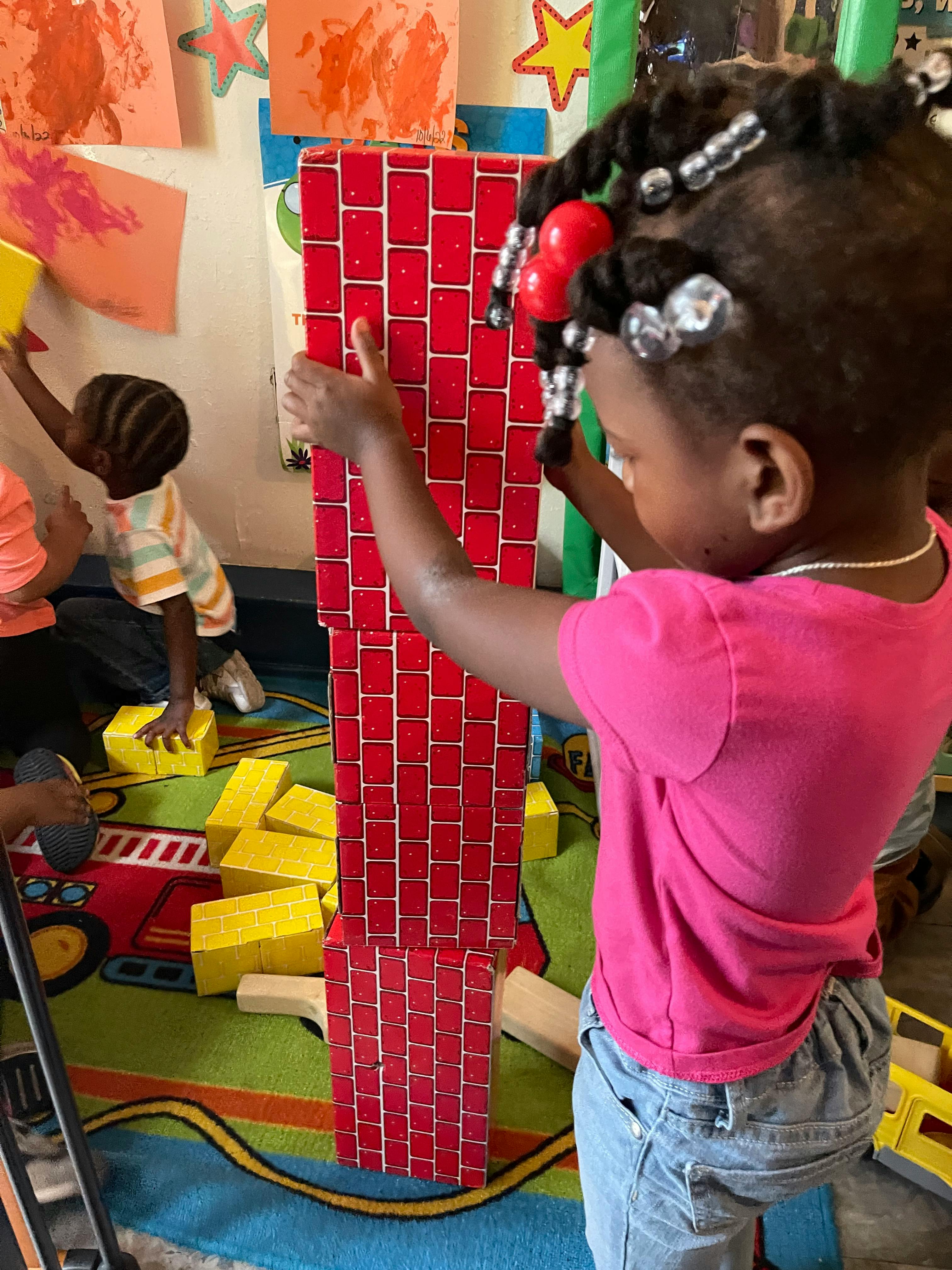 Child building a tall structure with red and yellow blocks in a classroom