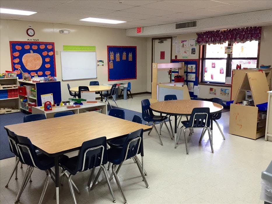 Empty classroom with small tables and chairs, shelves with toys, and an art wall