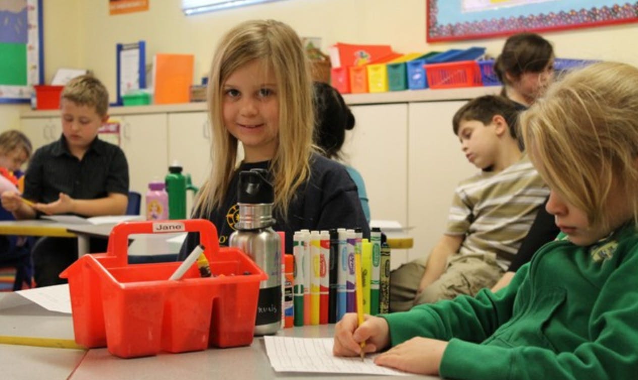Child writing at a desk in a classroom with other children