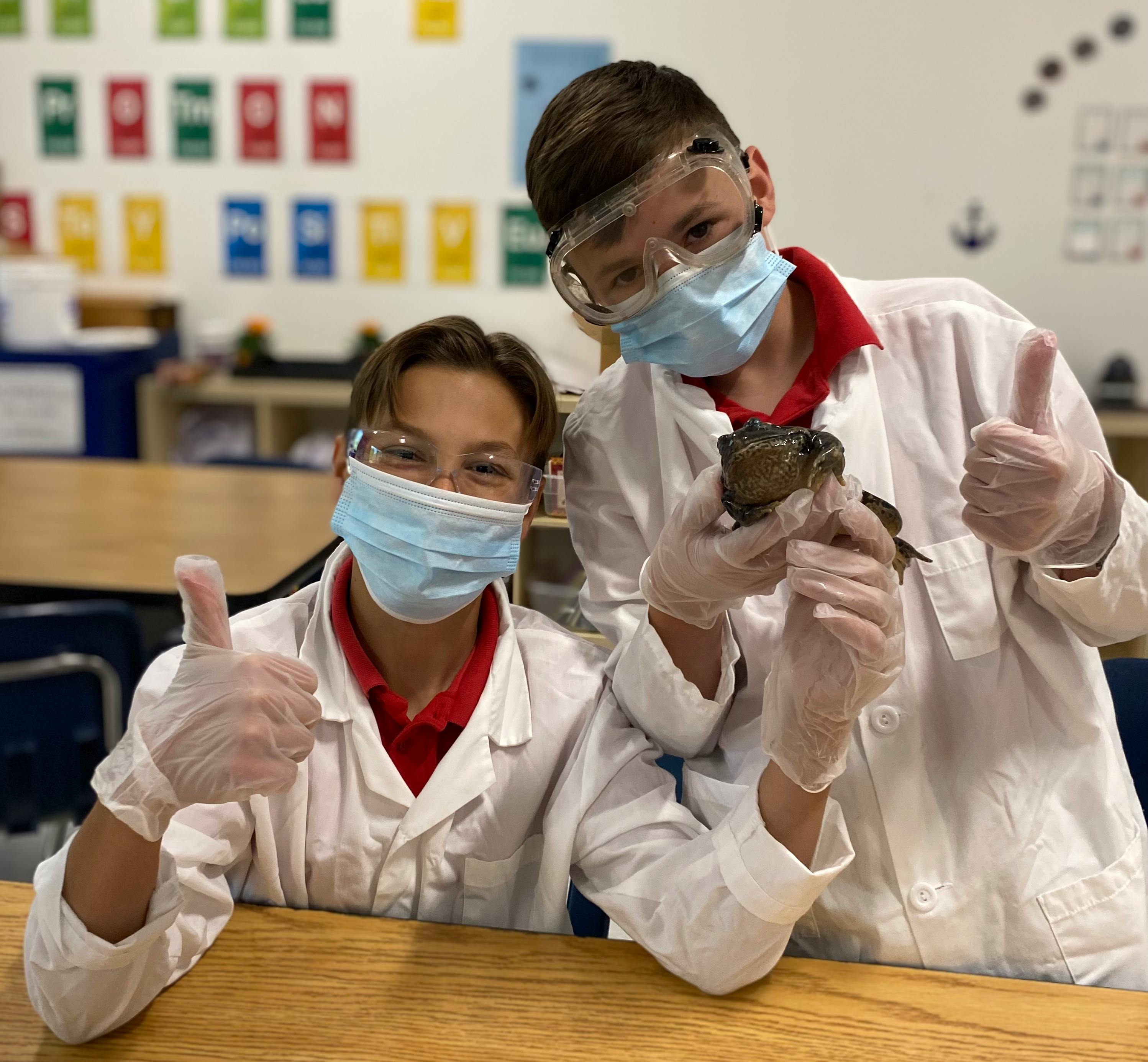 Two teens in lab coats, masks, and gloves, one holding a frog, both giving thumbs up