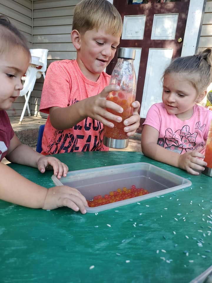 Three children at a table, one holding a bottle with liquid and beads