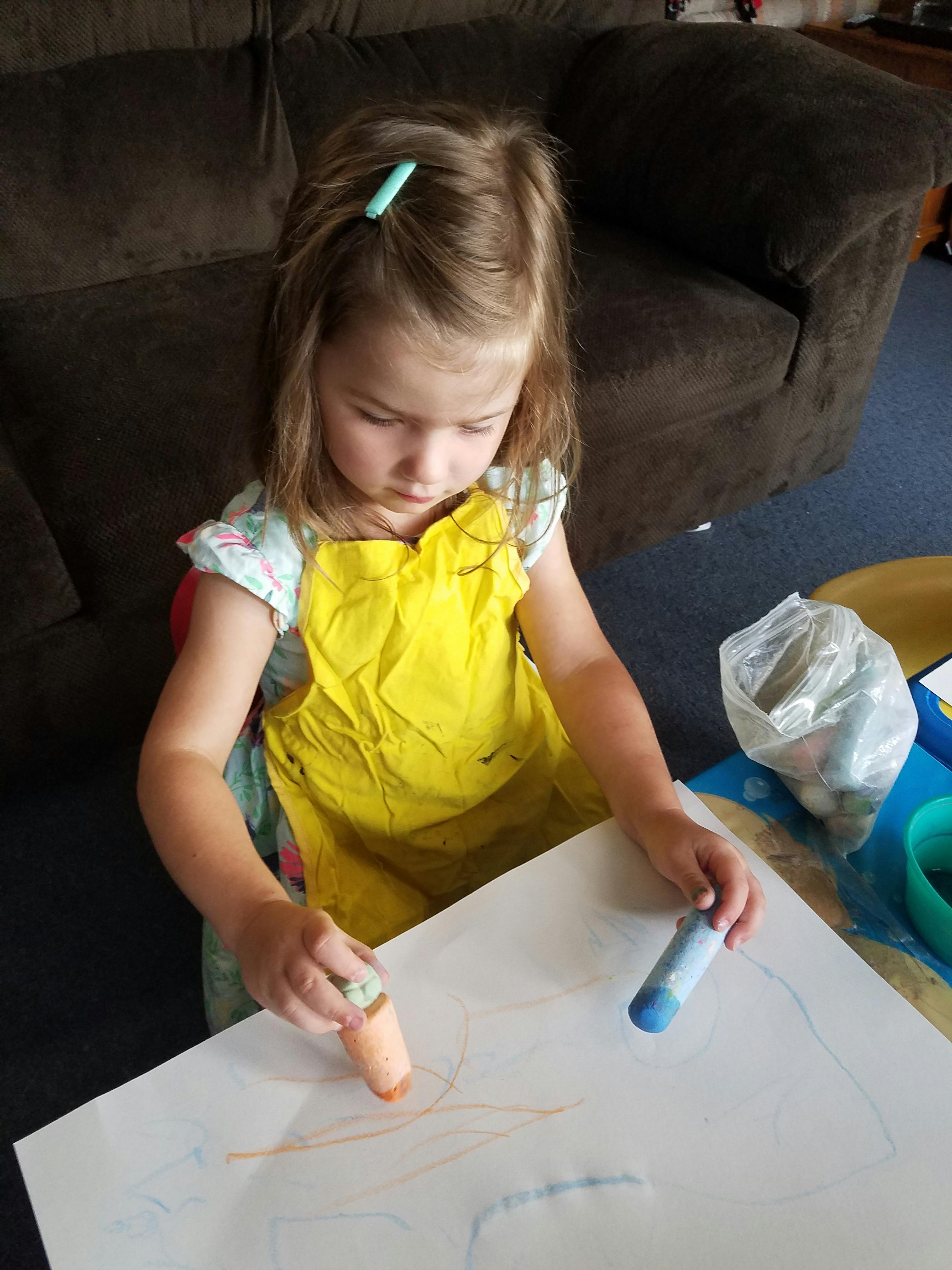 Child in yellow apron drawing on paper with crayons