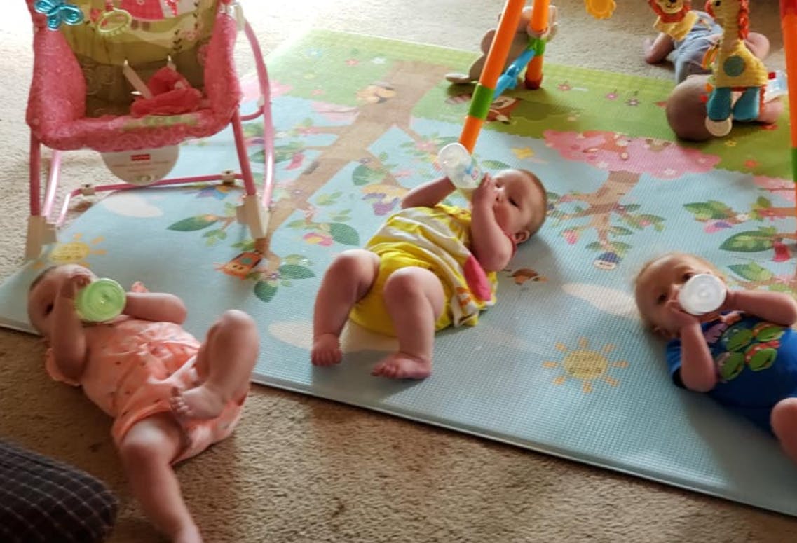 Three babies lying on a playmat drinking from bottles