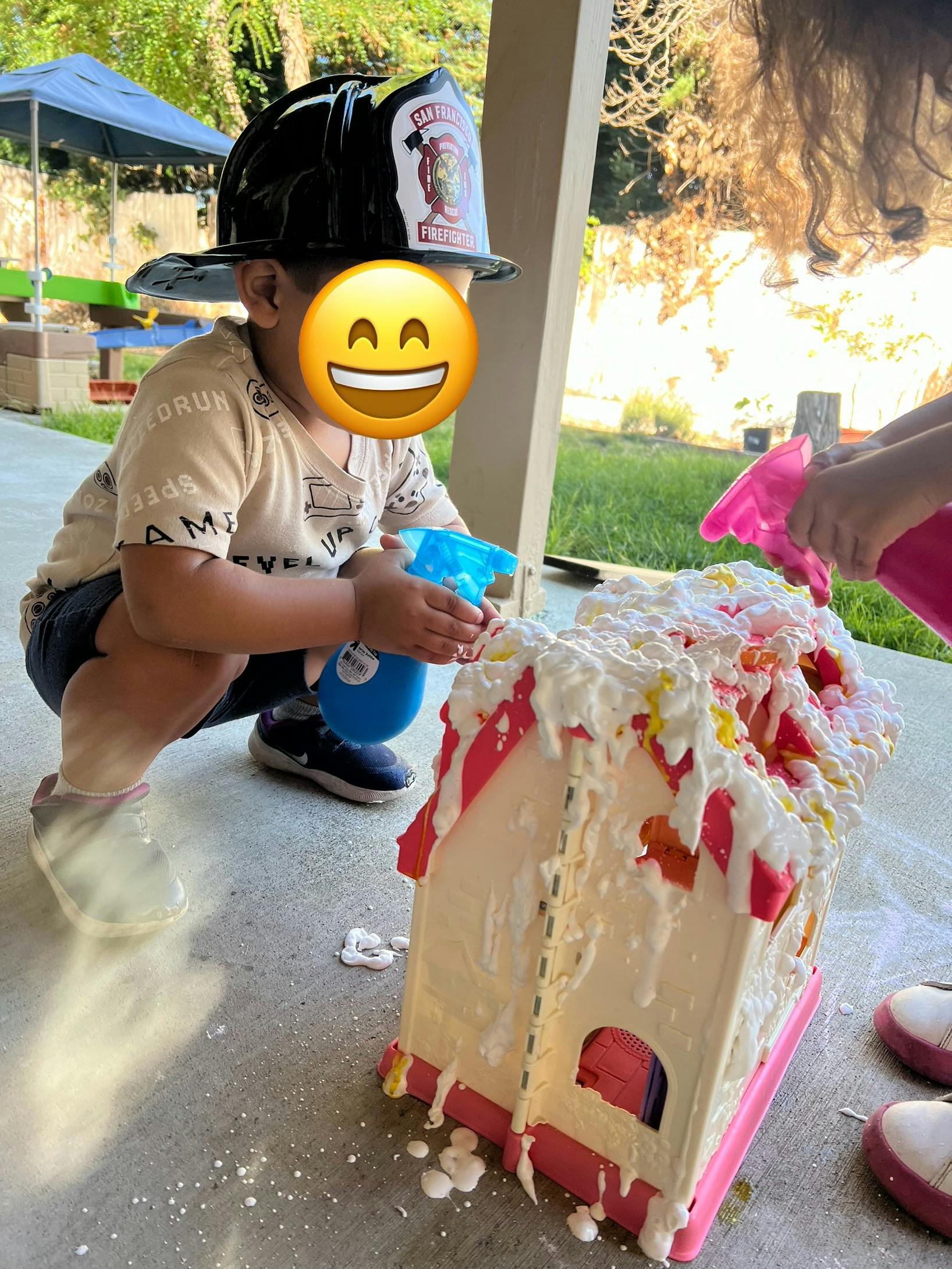 Child in firefighter hat spraying a toy house covered in foam