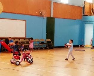 Child playing with a ball in a gym, another child on a toy scooter