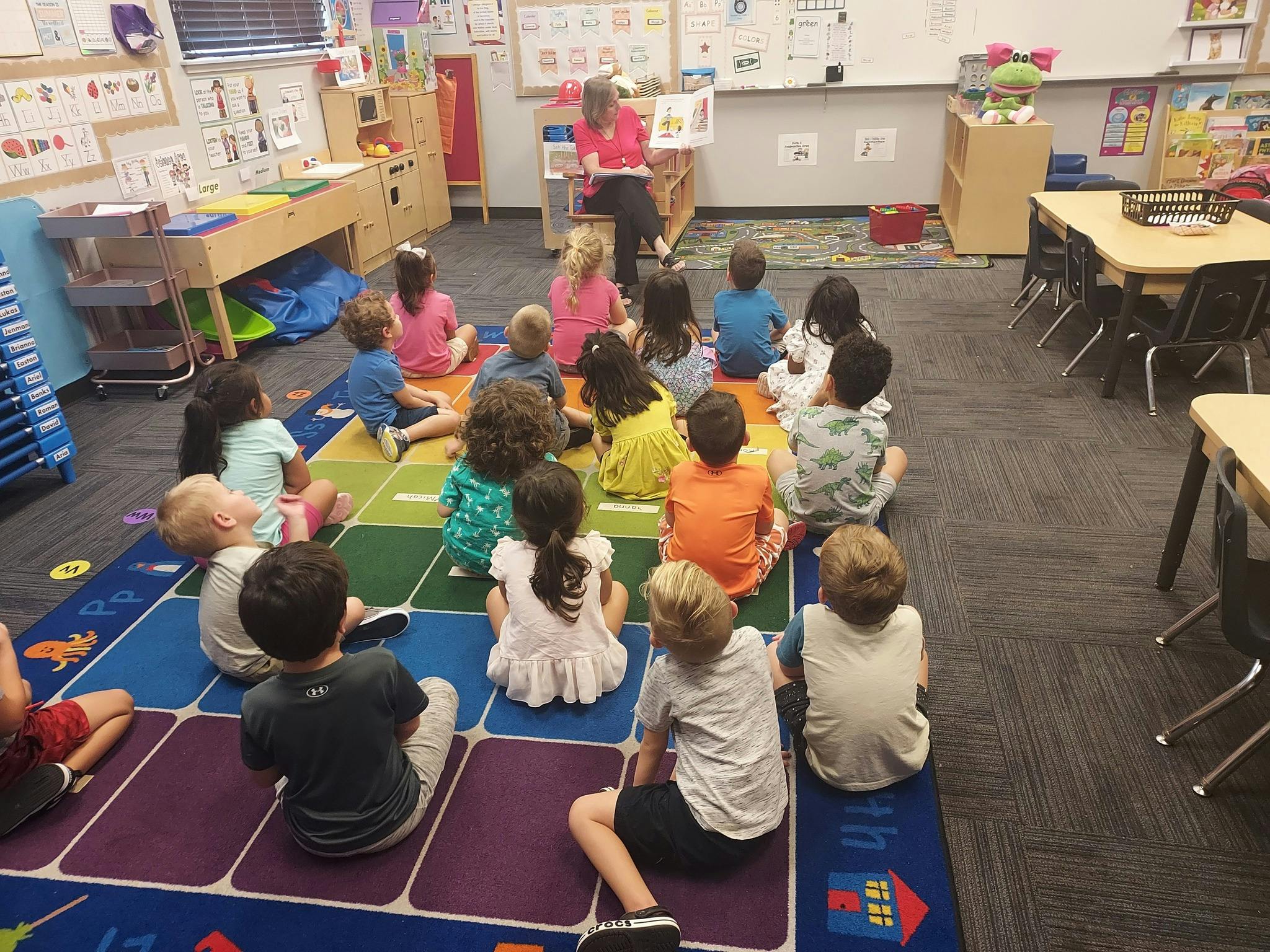 Person reading to a group of children sitting on a colorful rug in a classroom