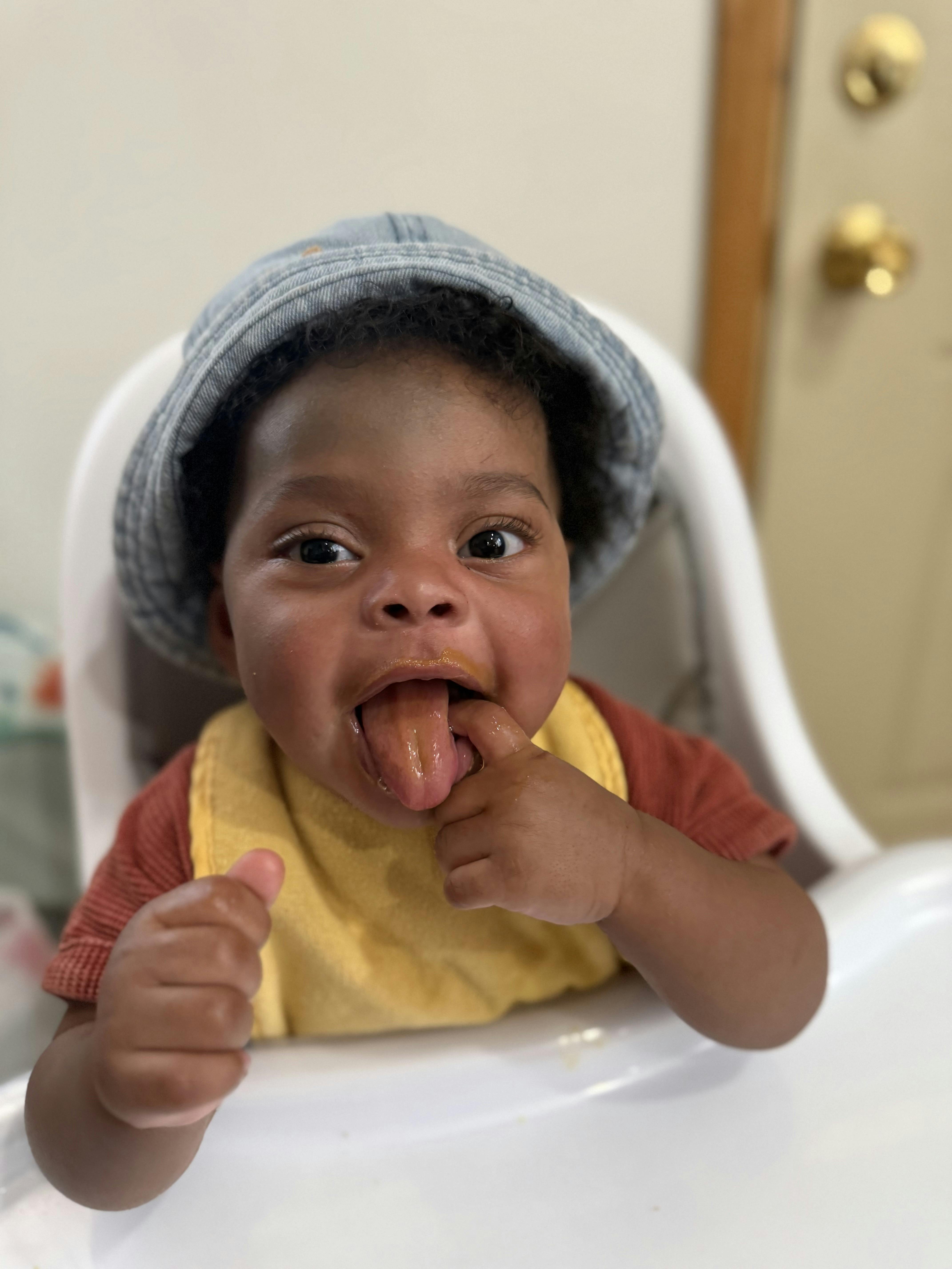 Child wearing a hat in a high chair with a finger in their mouth and tongue sticking out