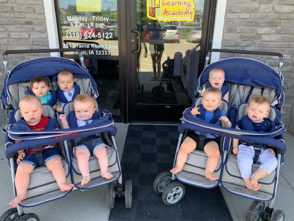 Six babies in two double strollers outside a building