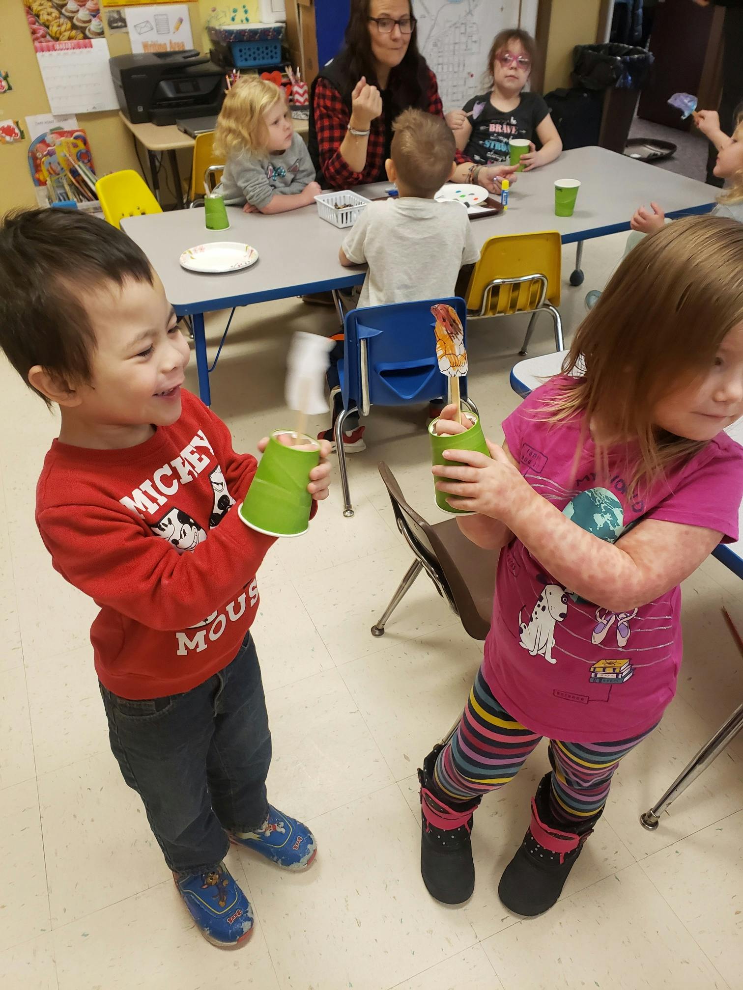Two children holding green cups, with other children and an adult in the background