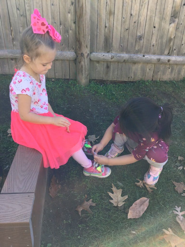 Child tying the shoe of another child sitting on a bench
