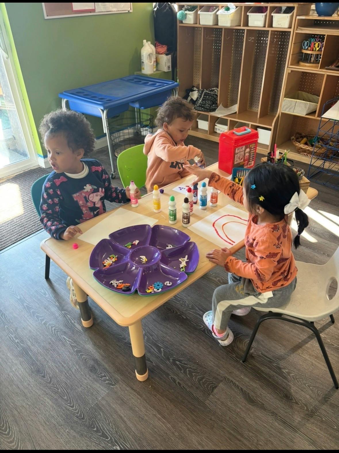 Three children sitting at a table using dot markers for arts and crafts