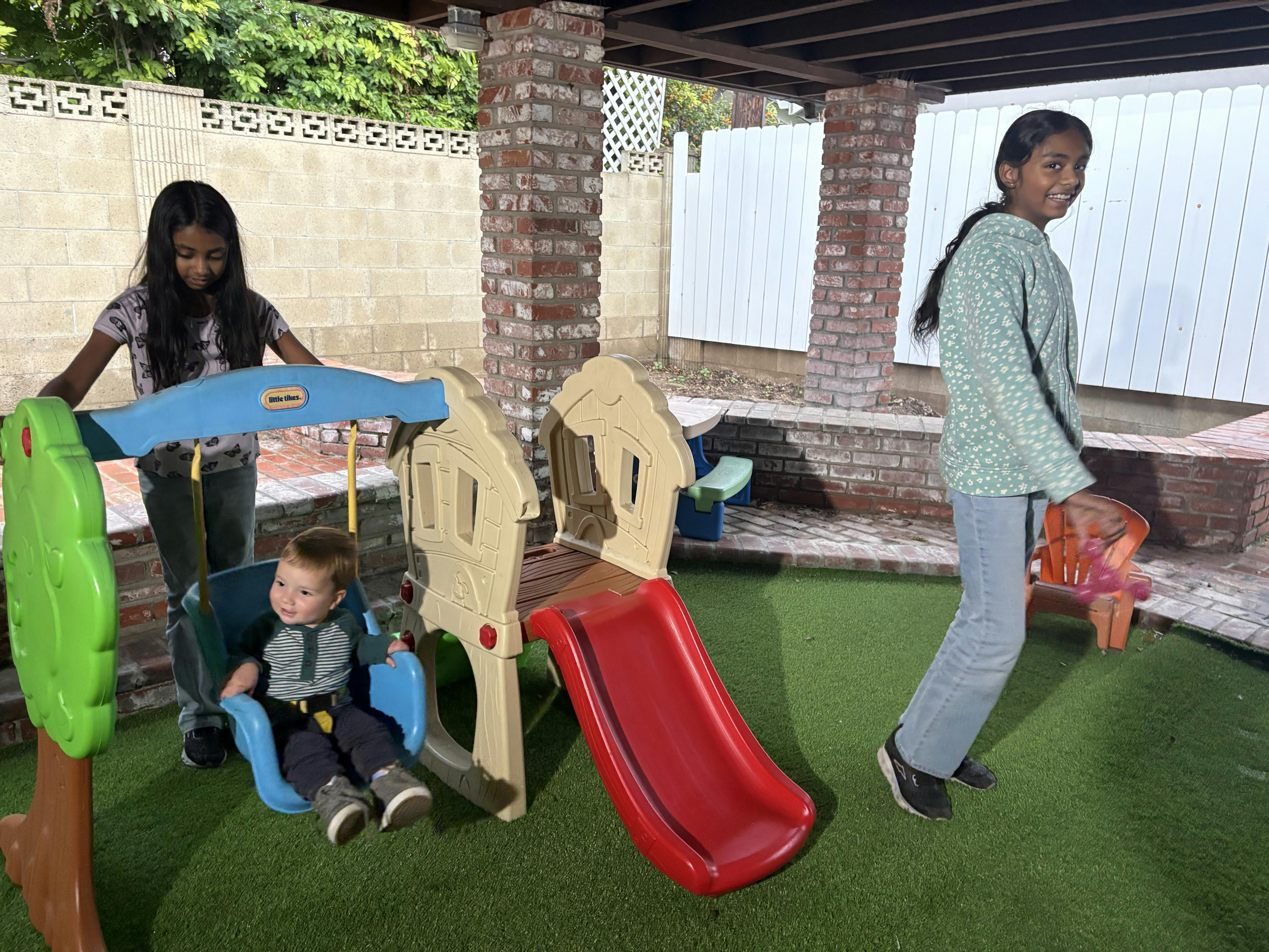 Child sitting in a swing next to a play structure with a slide, two other children nearby