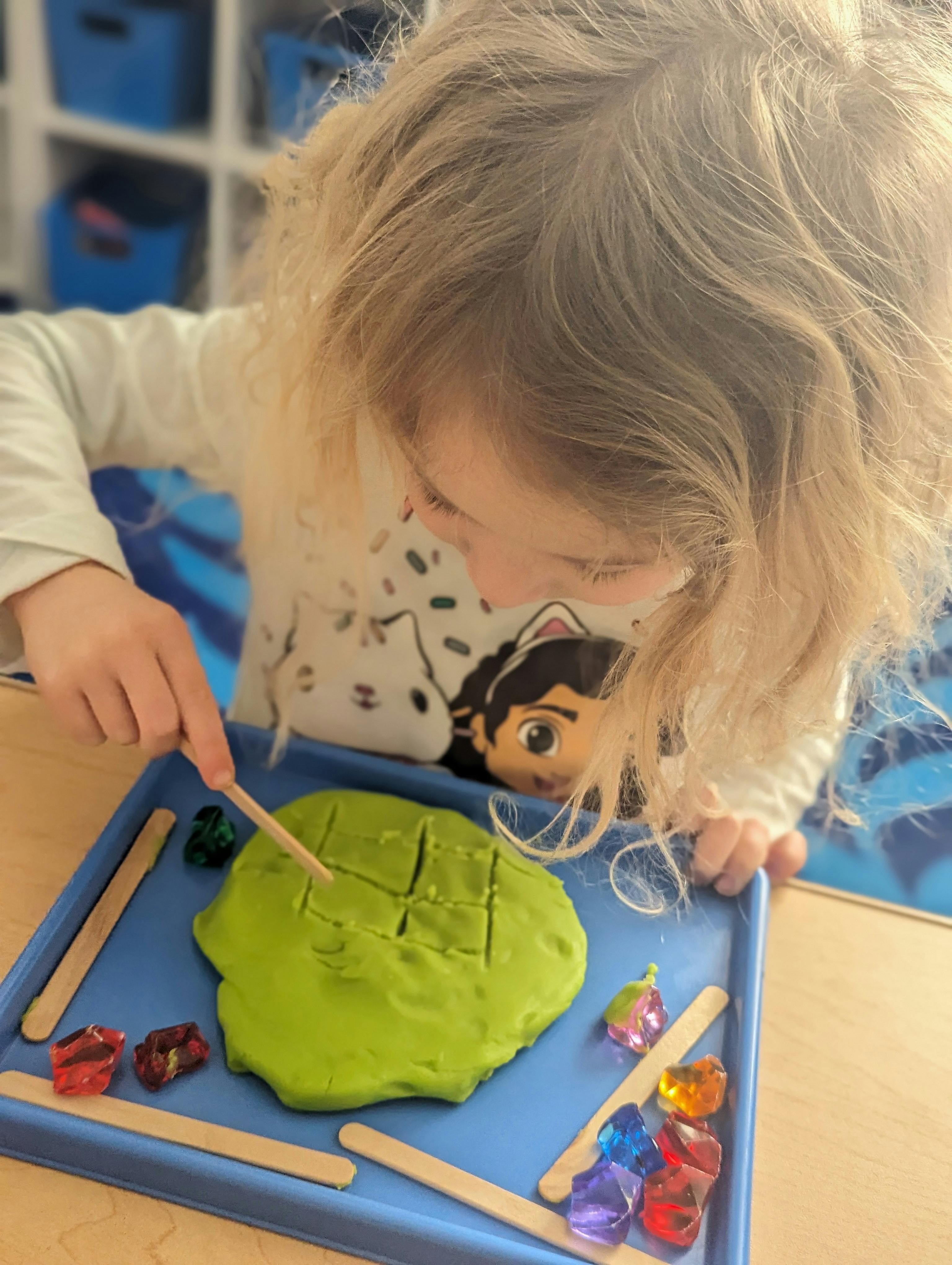 Child playing with green playdough and craft sticks on a blue tray