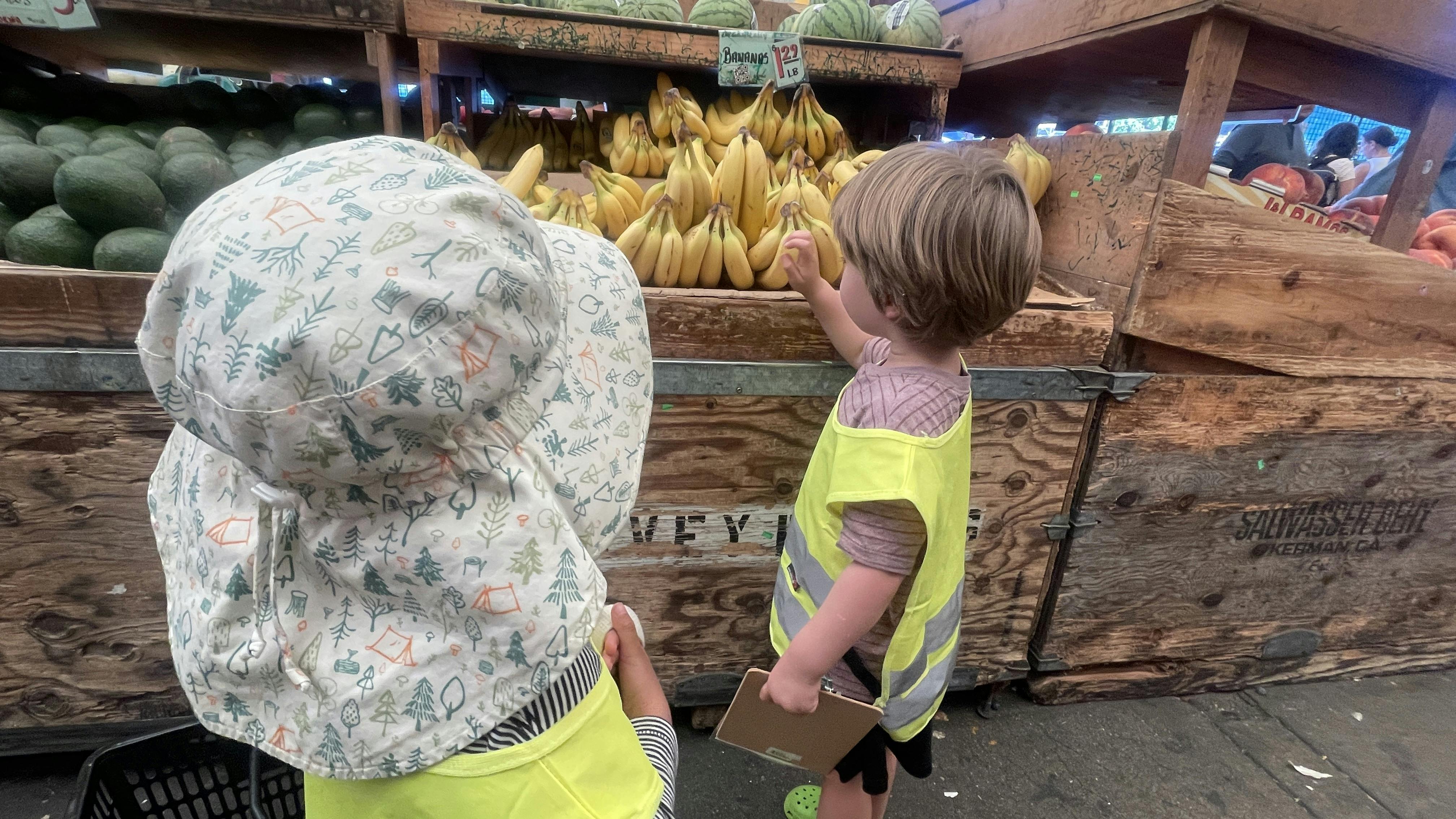 Two children wearing neon safety vests choosing bananas at a market
