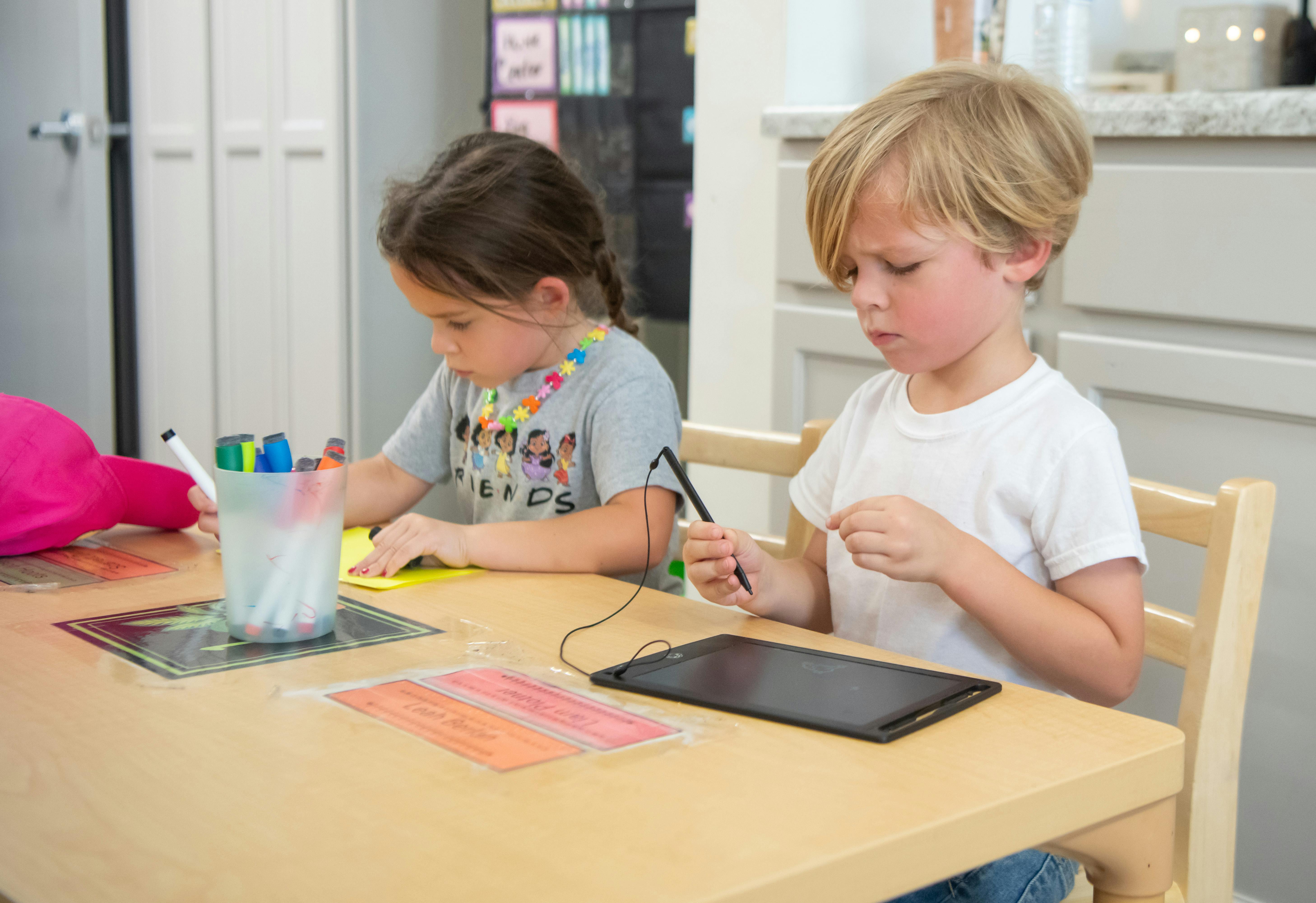 Two children at a table, one drawing and one holding a stylus