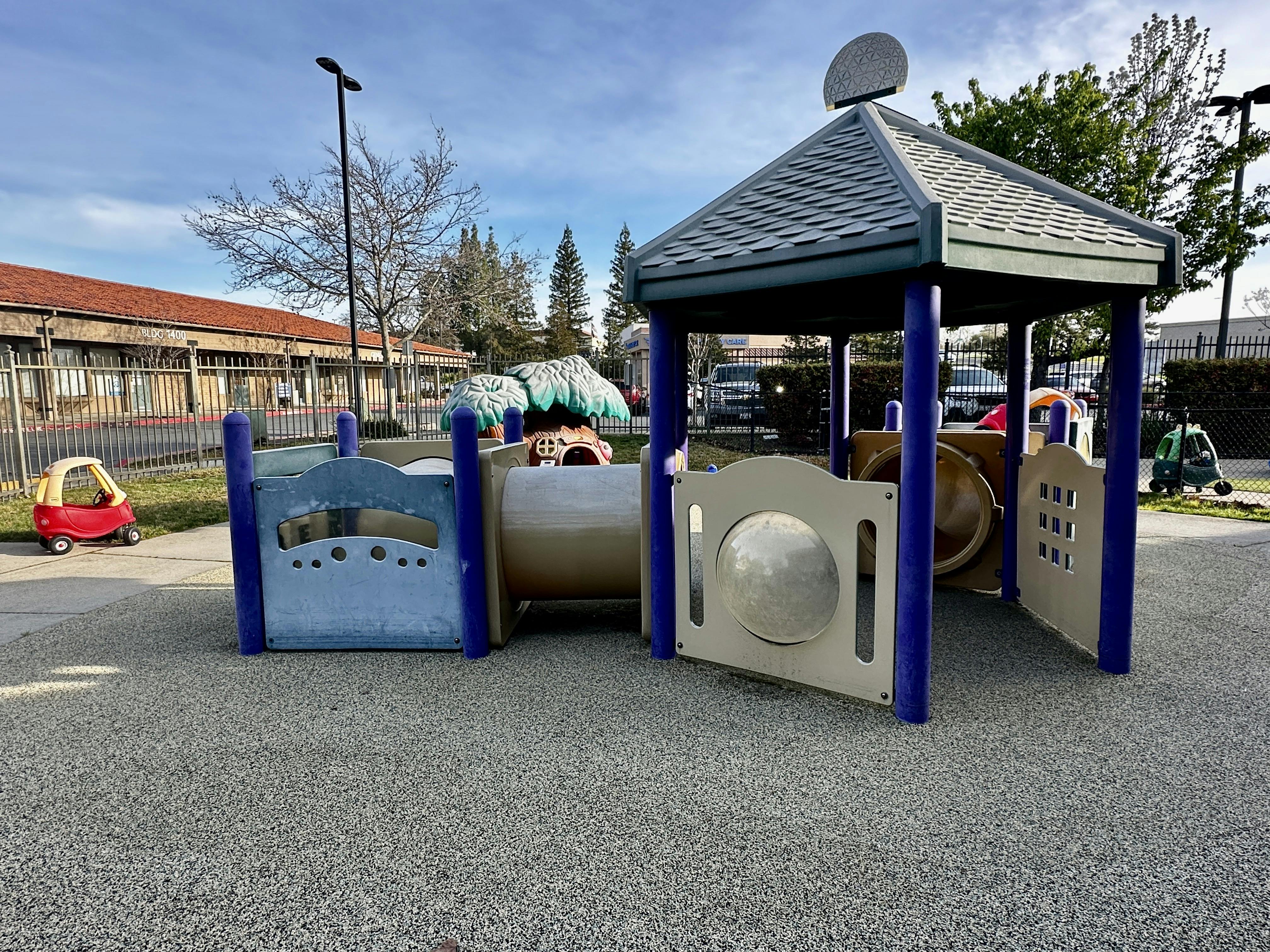 A covered play structure with a tunnel and an empty red toy car on a playground