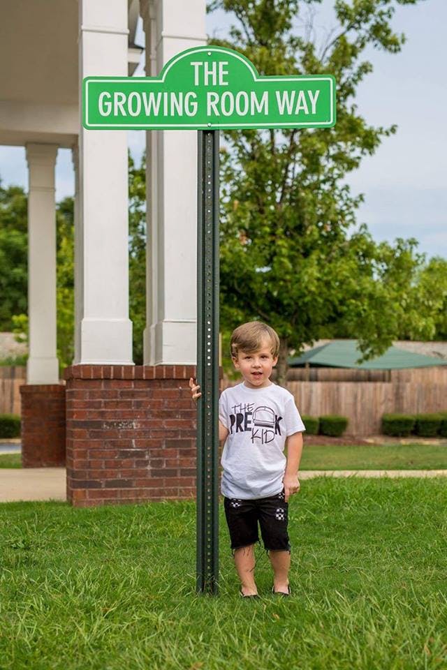Child standing next to a street sign that reads "The Growing Room Way"