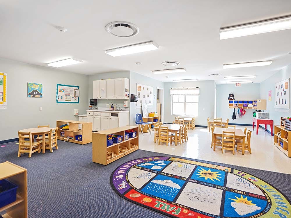 An empty classroom with small wooden tables and chairs, a sink, and a circular rug