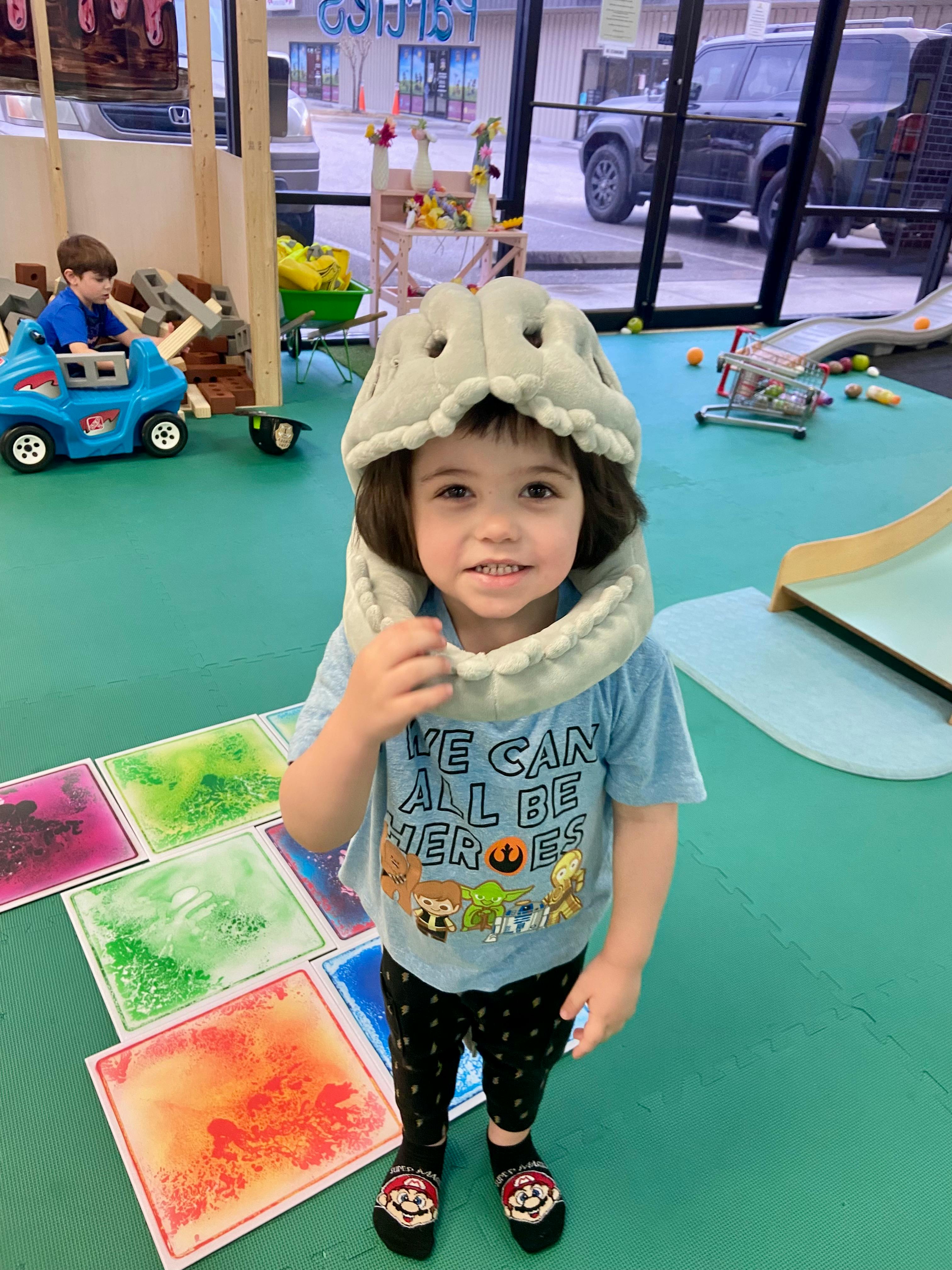 Child wearing a dinosaur hat and a shirt that says "WE CAN ALL BE HEROES"