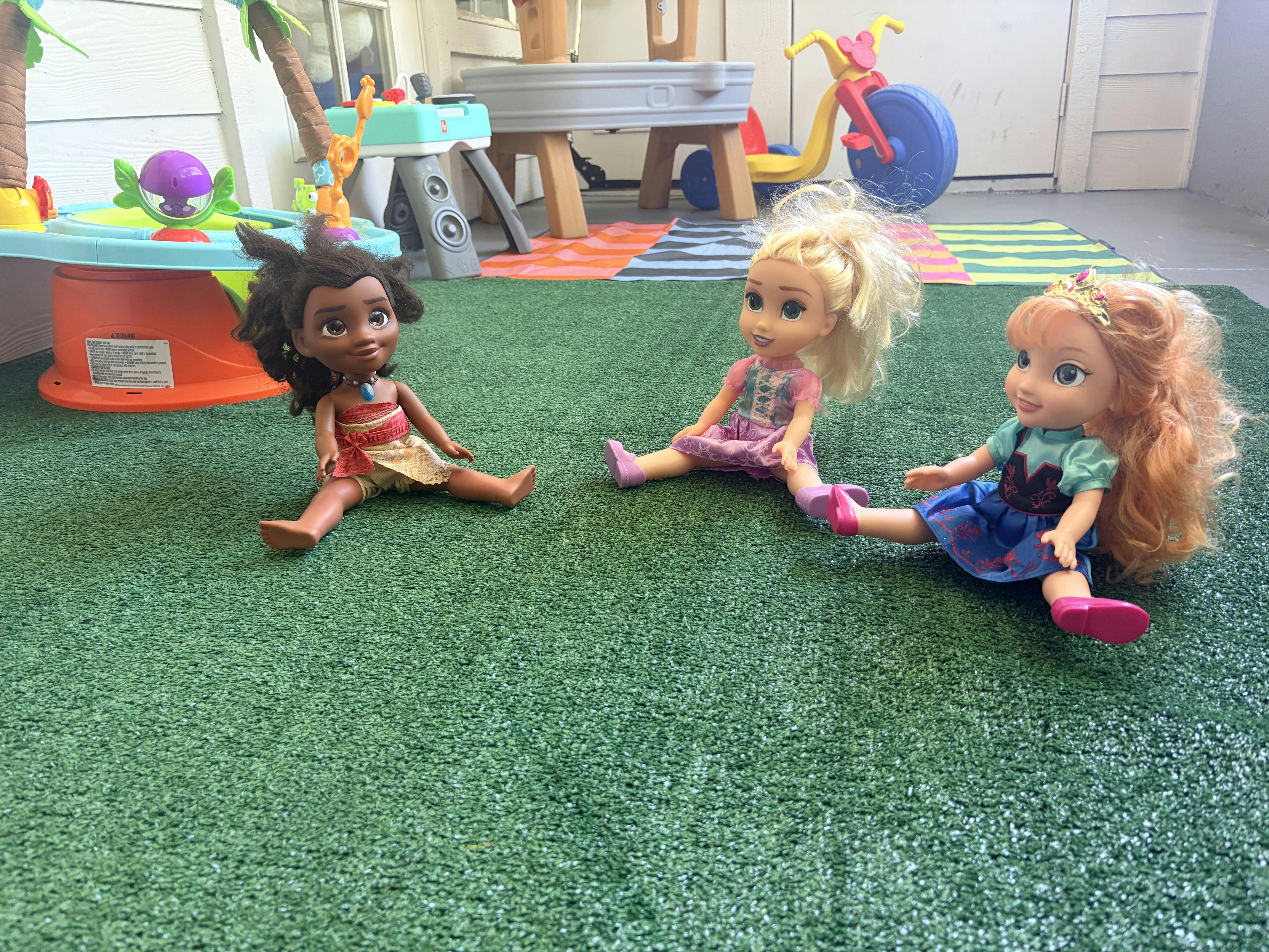 Three dolls sitting on a green surface with various toys in the background