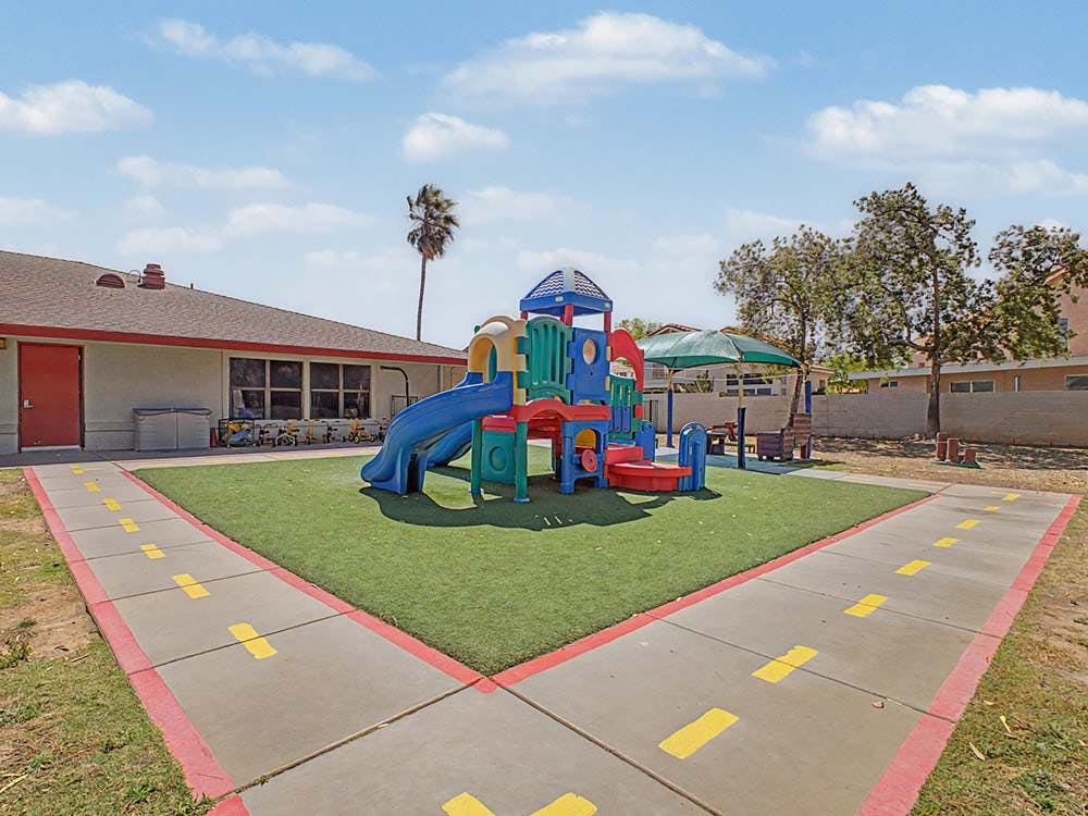 Colorful playground structure on artificial turf with a building and palm tree in background